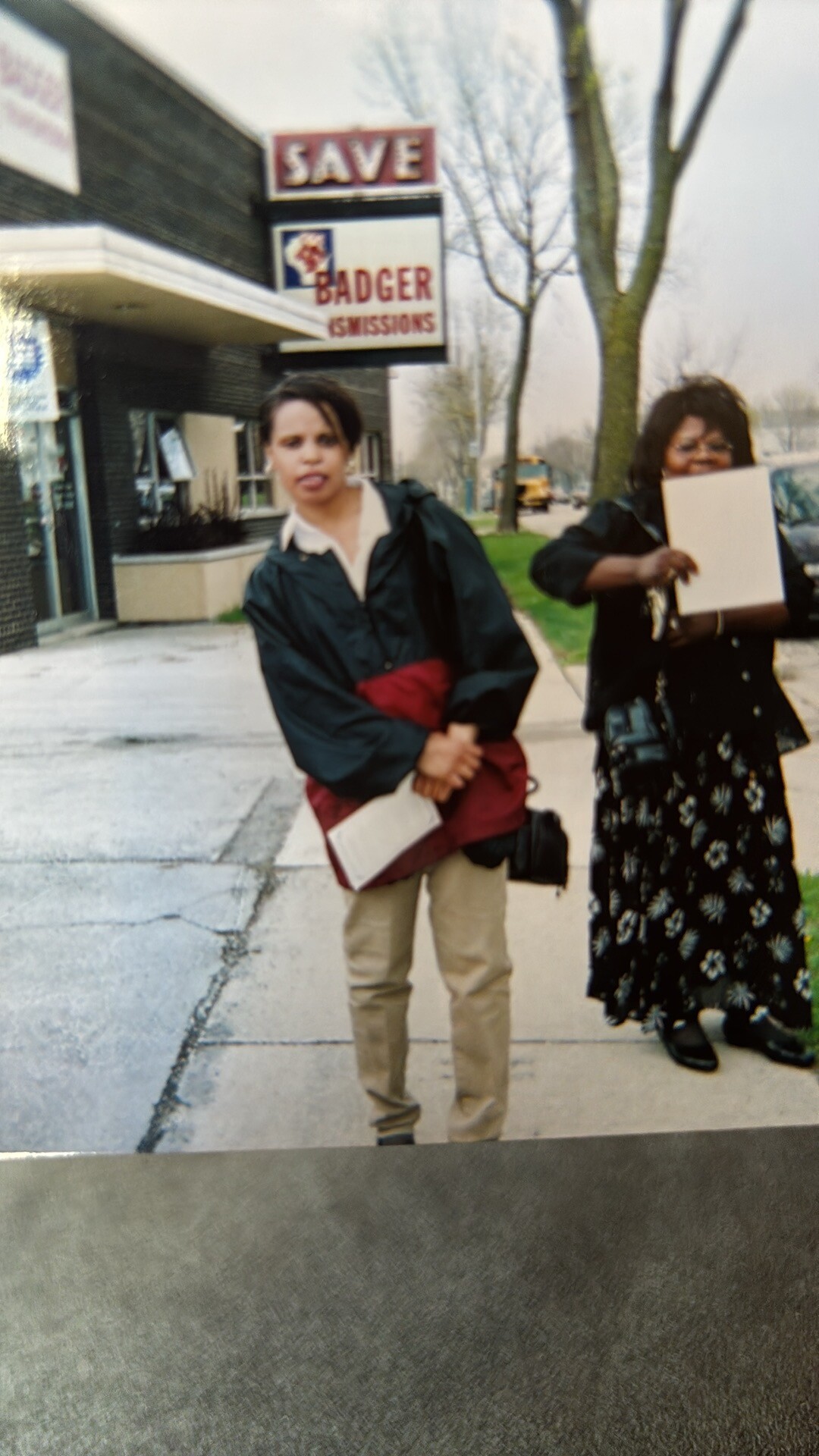 Two women walk on a sidewalk by a storefront with a 'SAVE BADGER MISSIONS' sign, one in a green jacket holding a red coat and papers, the other with a clipboard.