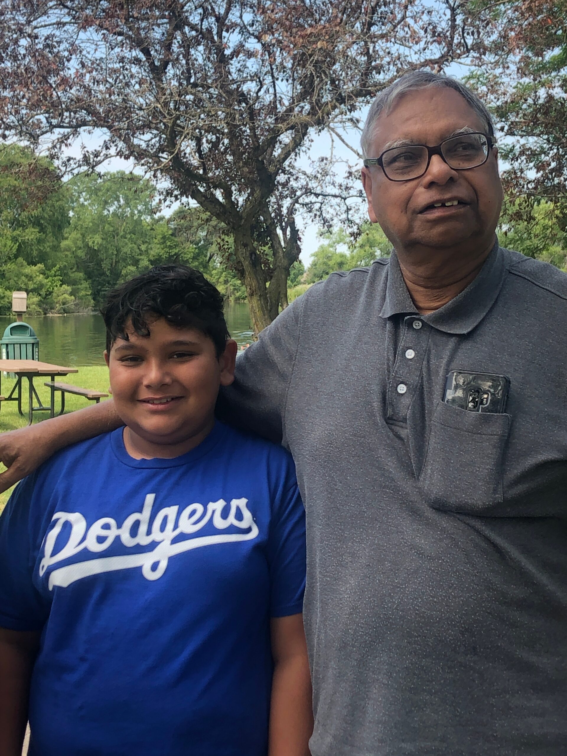 Two people, a man and a boy, pose together outdoors by a lake with trees in the background.