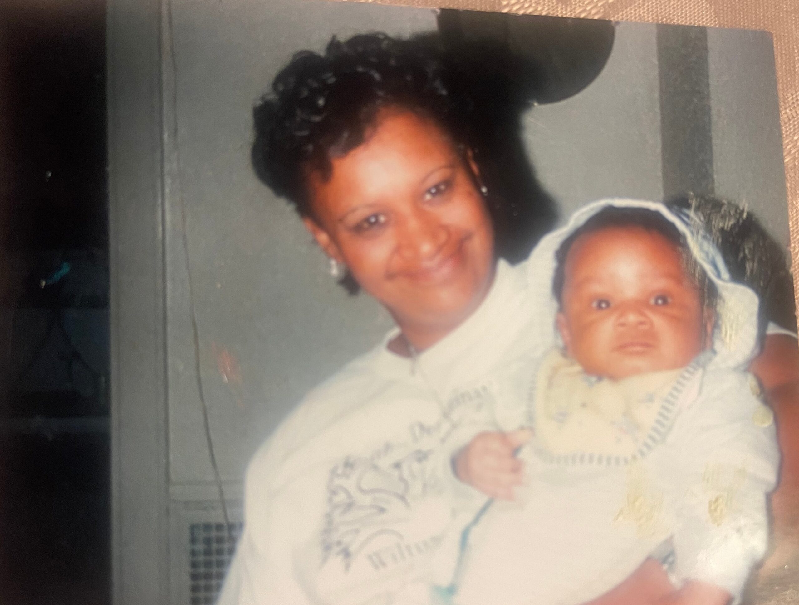 A smiling Black woman with short curly hair holding a baby, both wearing light clothing, in a photo on a scrapbook page.