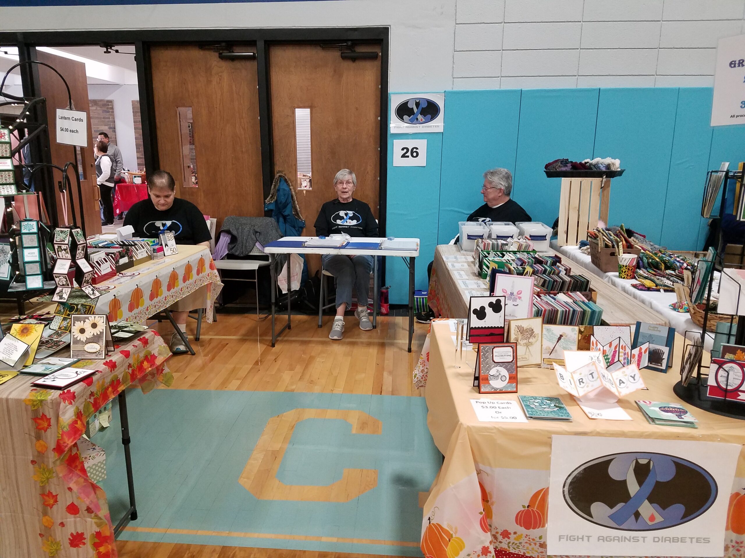Indoor craft fair with tables full of handmade cards and decorations; three people sit behind the tables at the back of the room.