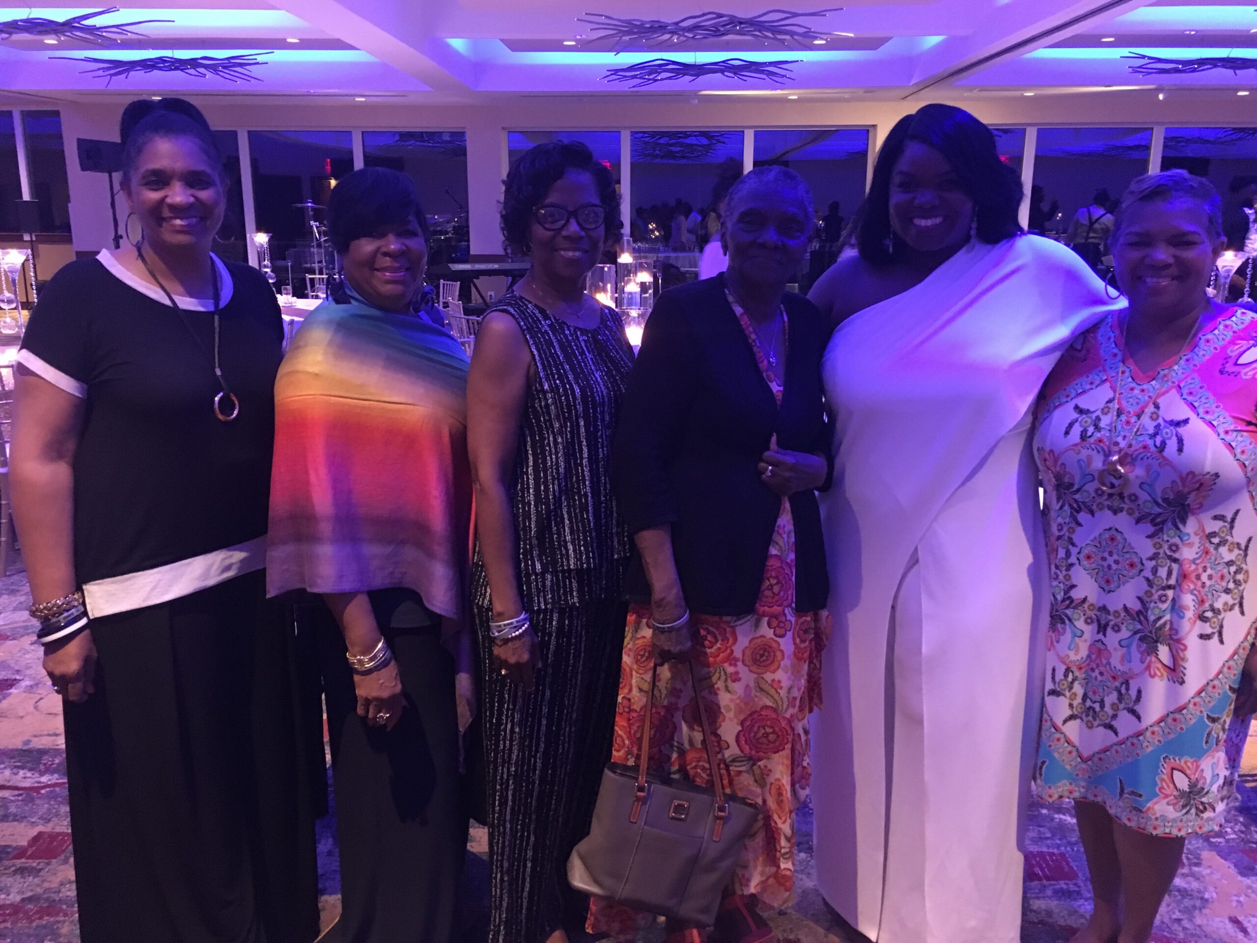 Six women posing together at a formal event, wearing colorful dresses, under blue lighting in a banquet hall.