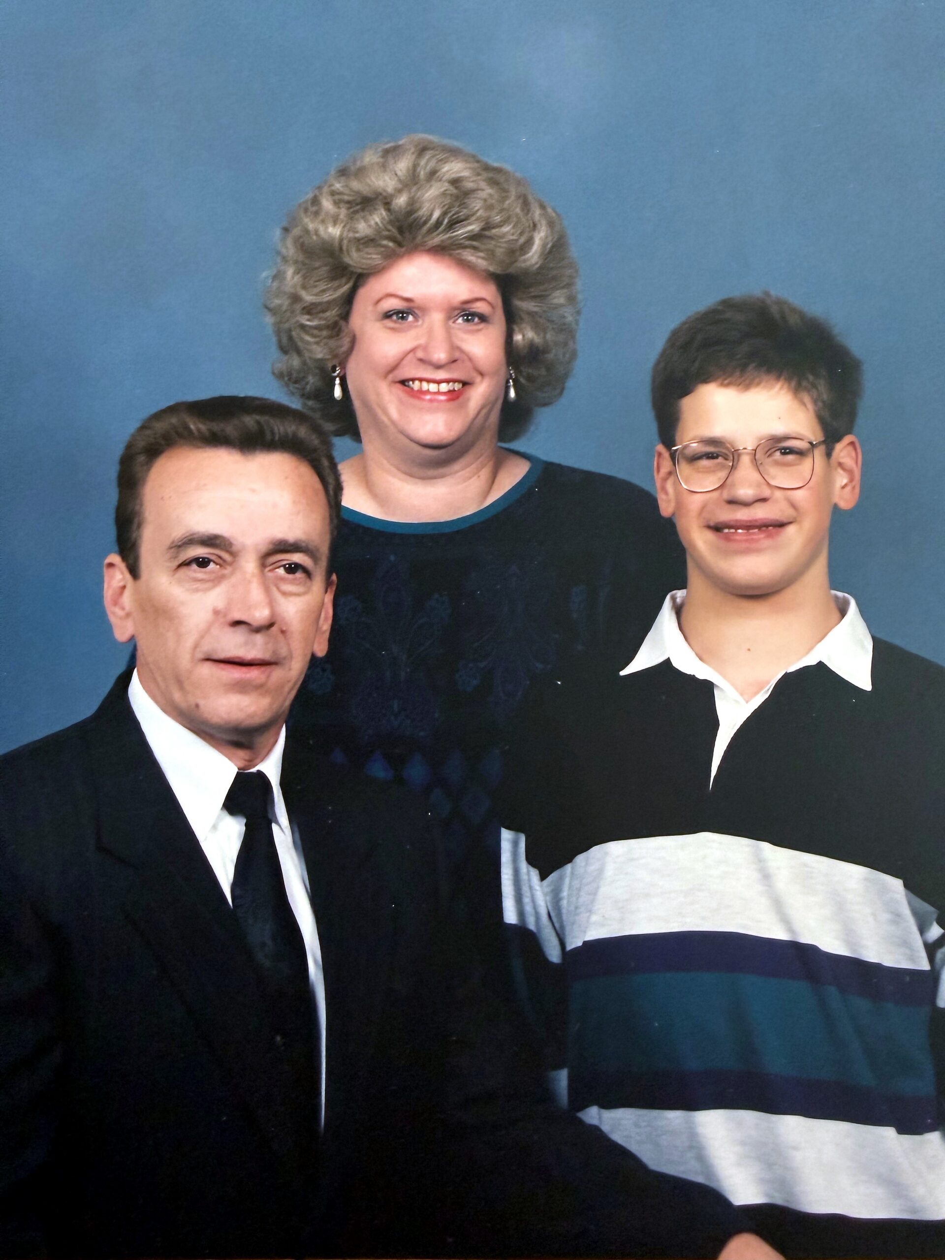 Portrait of a man, woman, and teenage boy posing together against a blue backdrop, smiling at the camera.