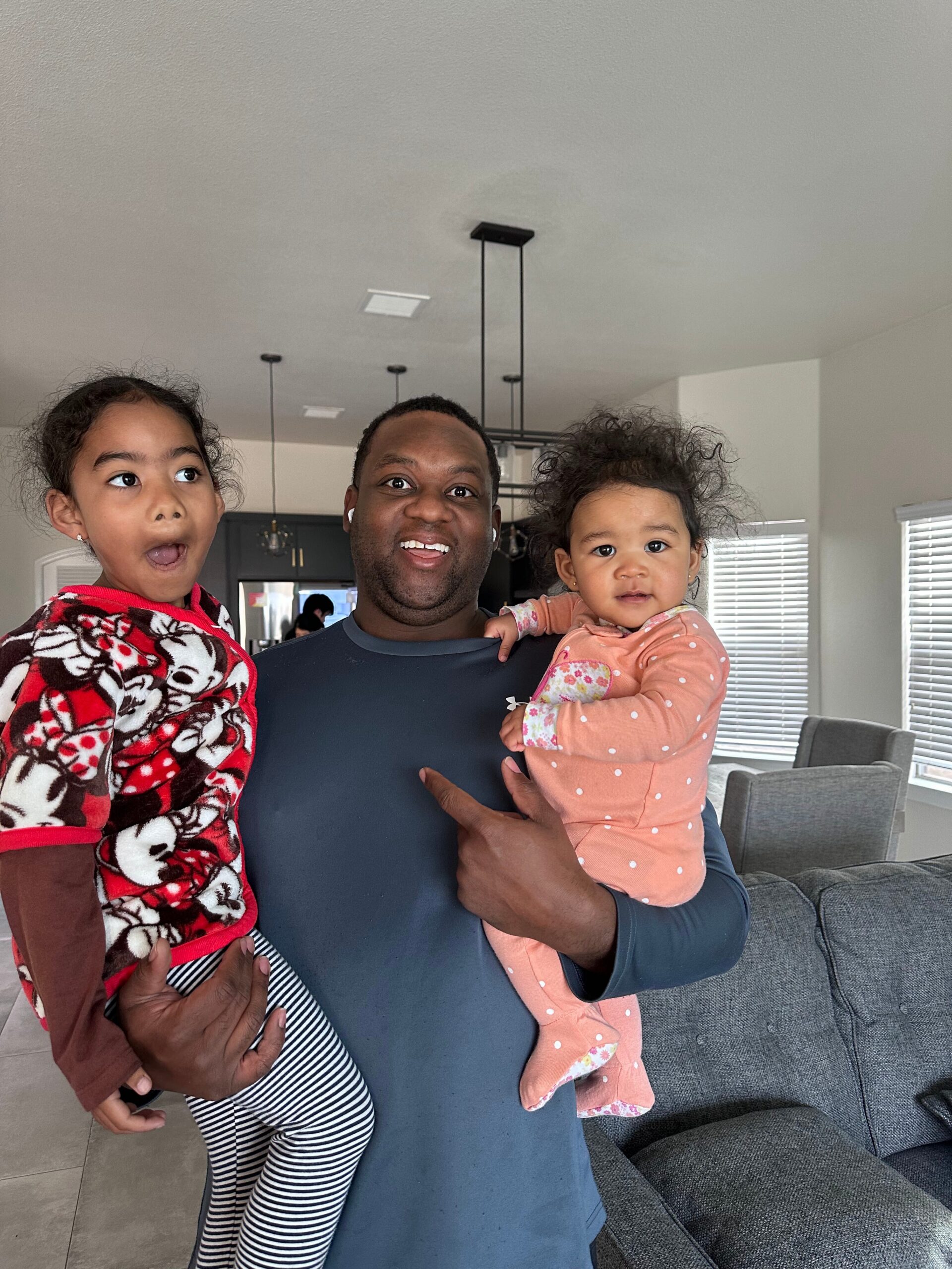 Man holding two young children indoors, one girl in a red Minnie Mouse fleece and the other in a peach polka-dot outfit, all smiling in a living room.