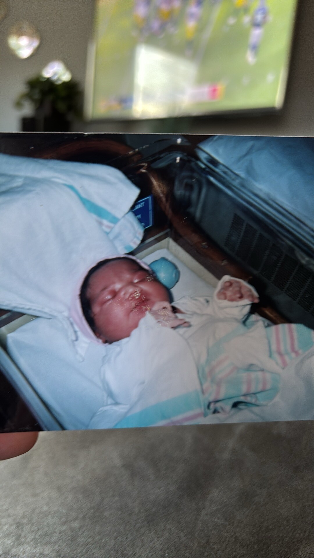 Newborn baby wrapped in blankets lying in a hospital bassinet, face slightly red with medical care nearby in a dim room