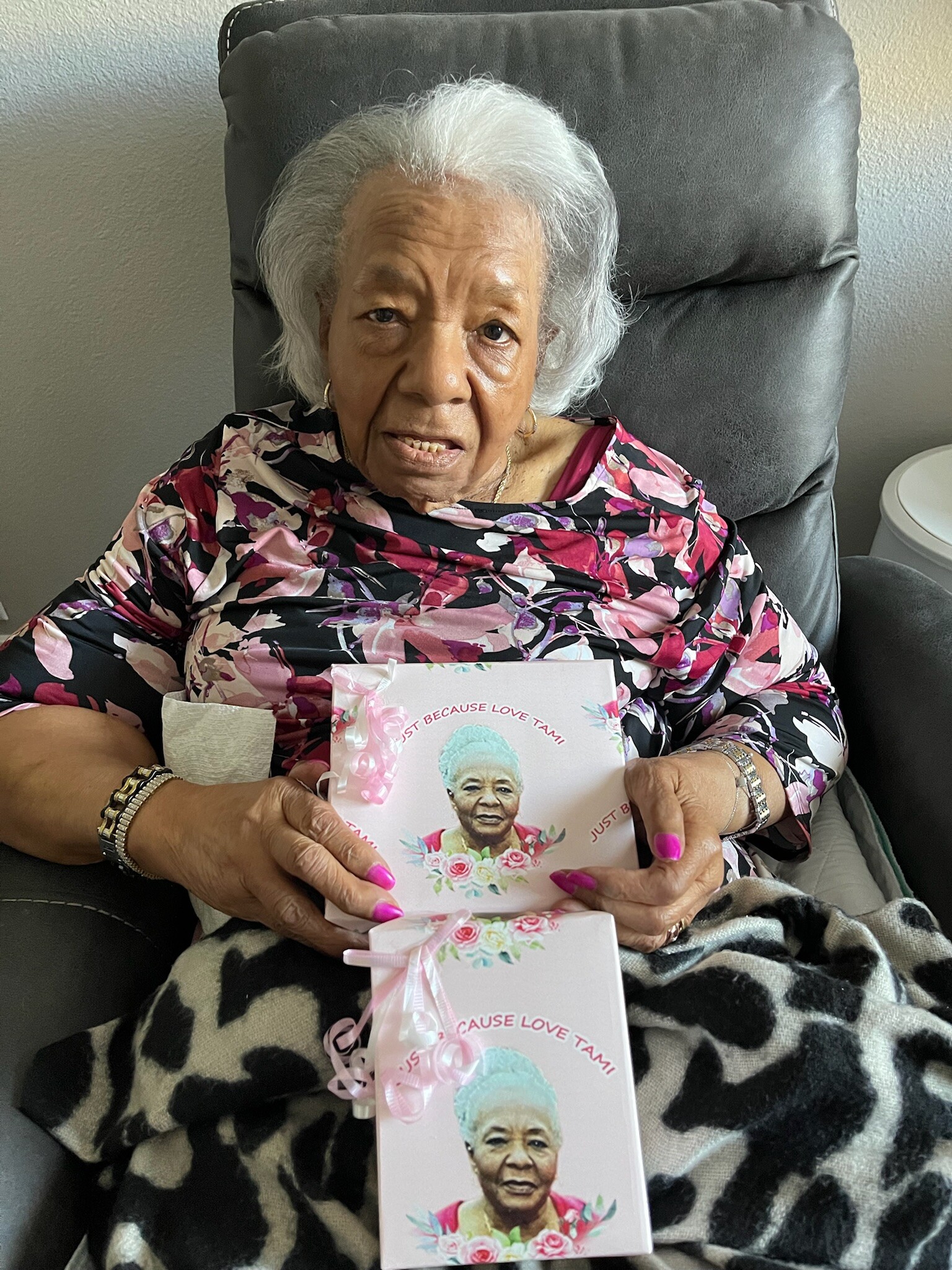 Older woman with white hair sits in a recliner, holding a pink gift card featuring her photo on the front.