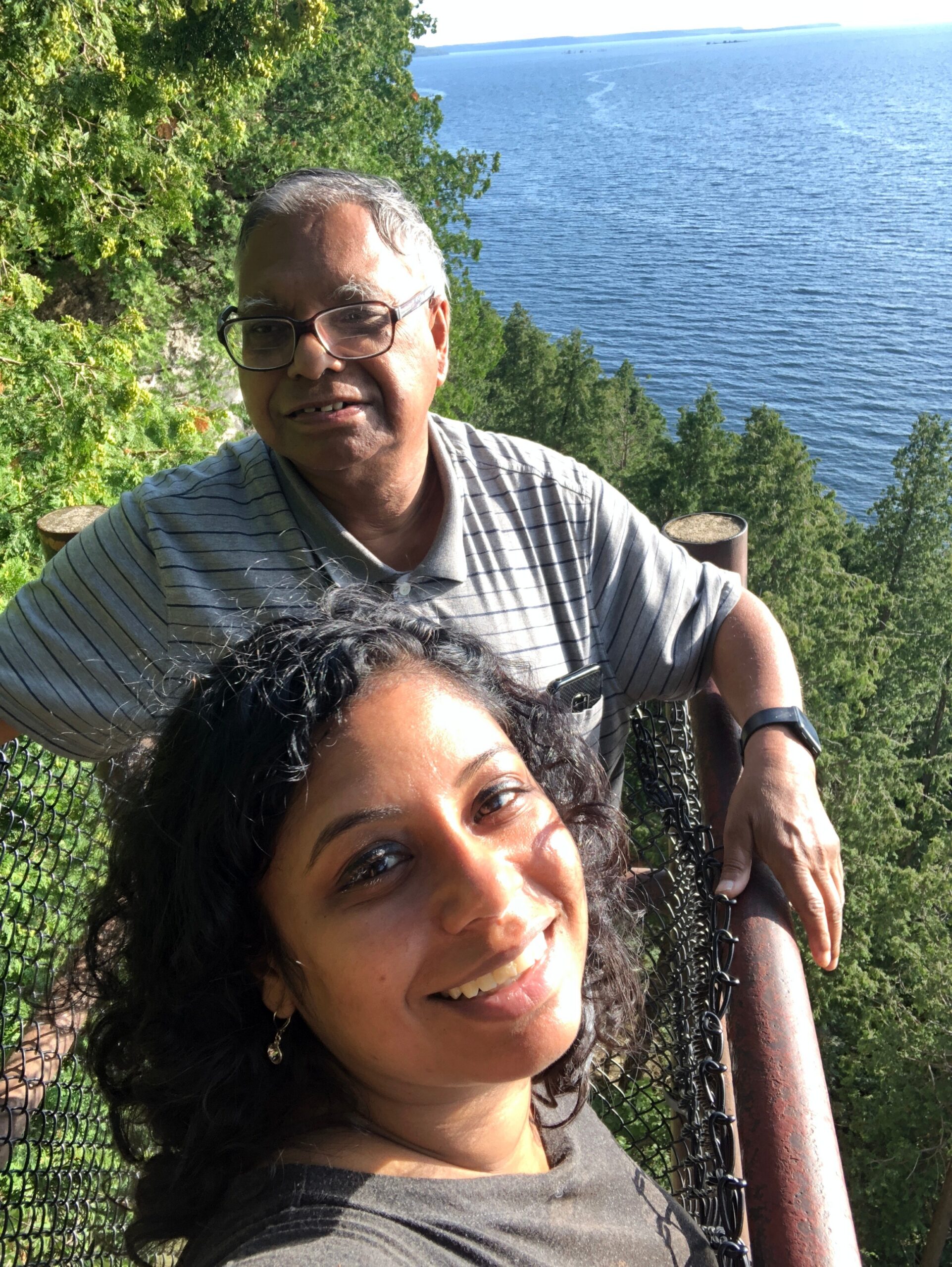 Smiling woman in the foreground with dark, curly hair and a man with glasses behind her, standing by a chain-link fence overlooking a blue lake and green trees.