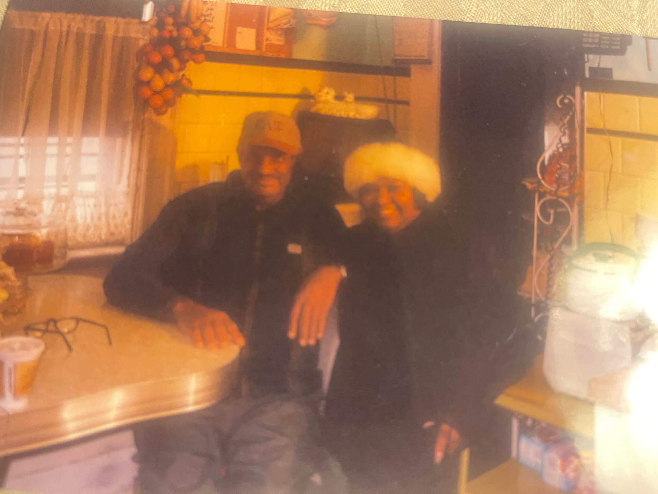 Two people wearing dark clothing pose for a warm, indoor photo on a cluttered table, with orange decorations visible in the background (pumpkins/balloons).