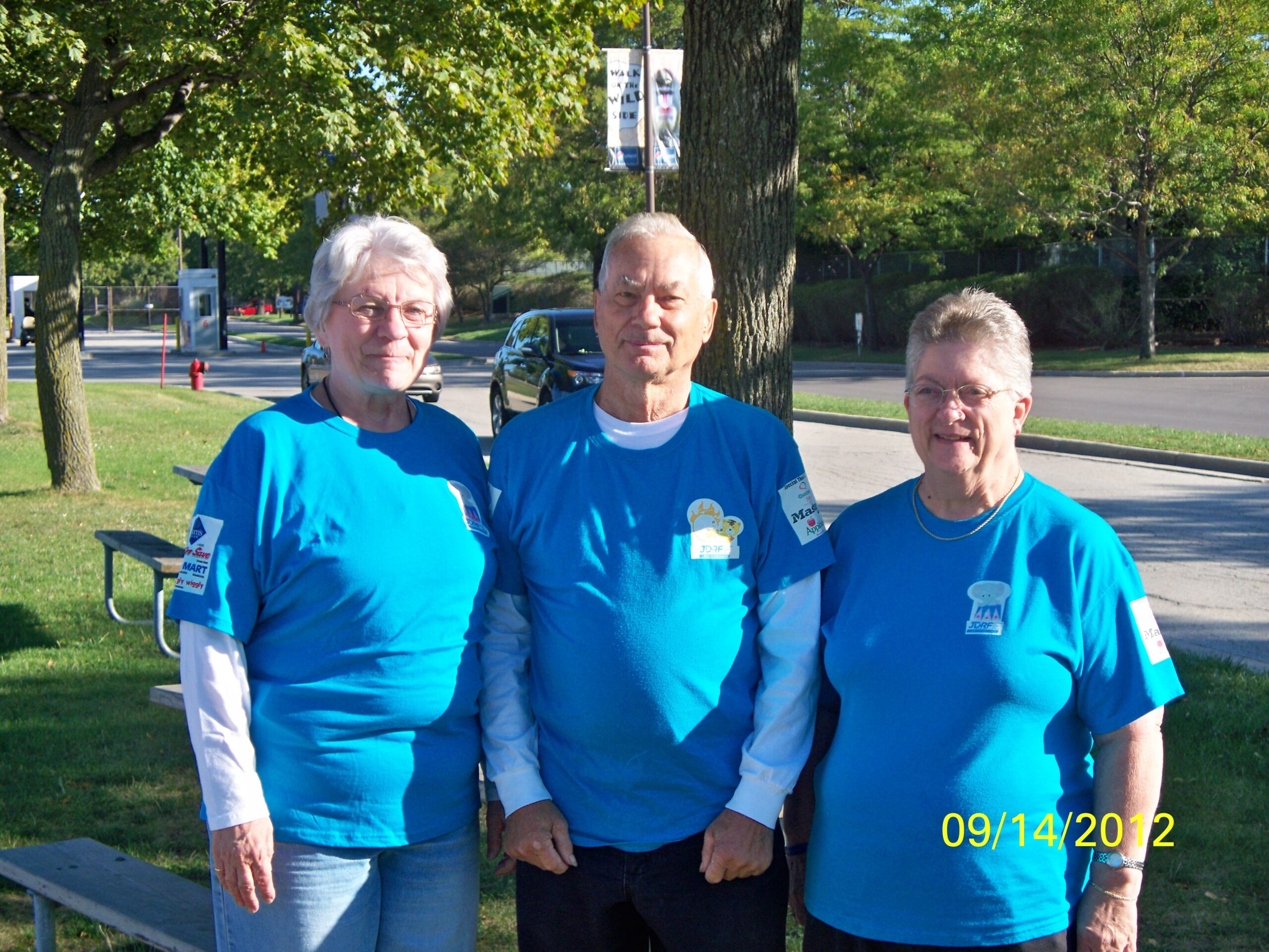Three adults in blue shirts posing together outdoors in a park with trees behind them.