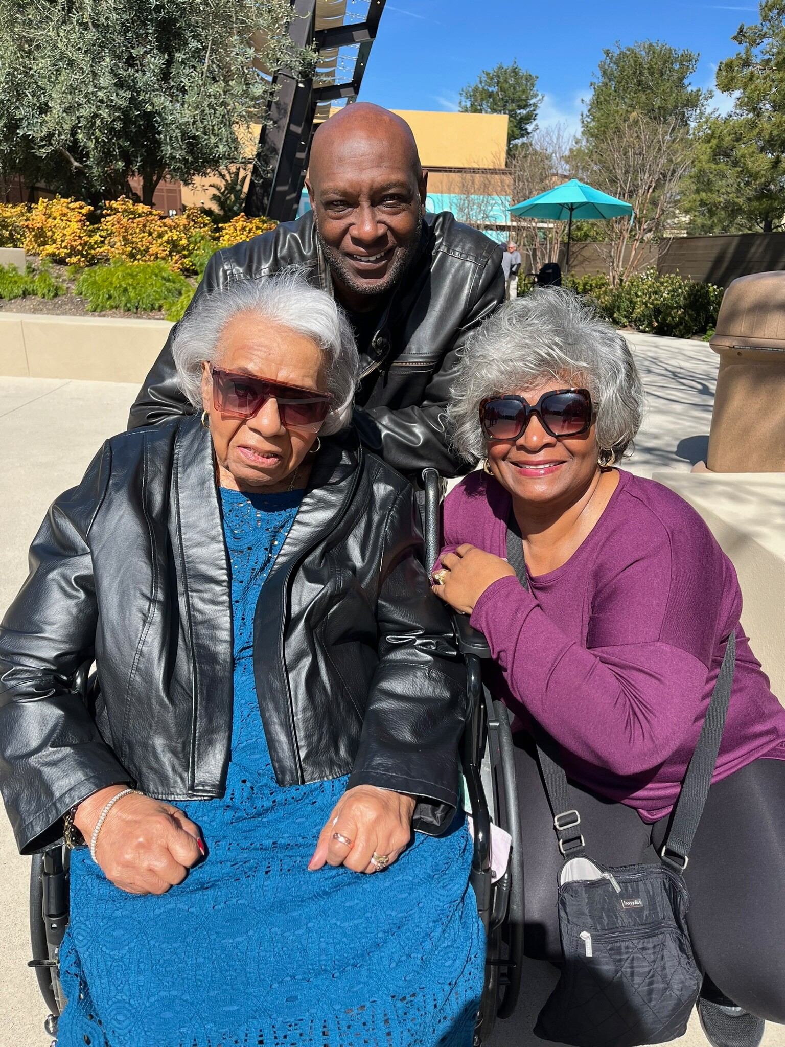 Smiling man leans over two elderly women in wheelchairs outdoors on a sunny day, with trees and a blue sky behind them.