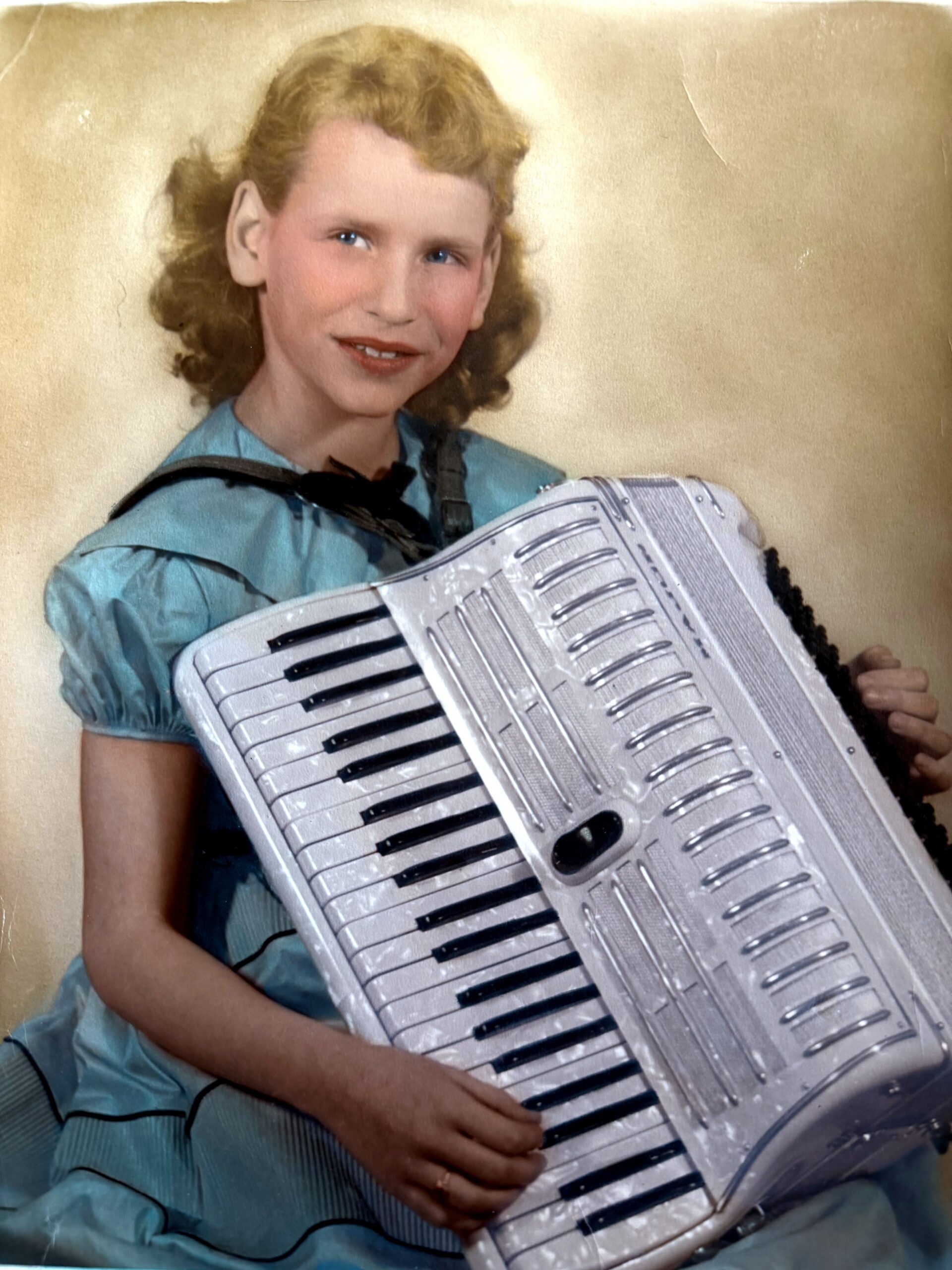 Young girl in a blue dress smiling while playing a silver accordion for a portrait.