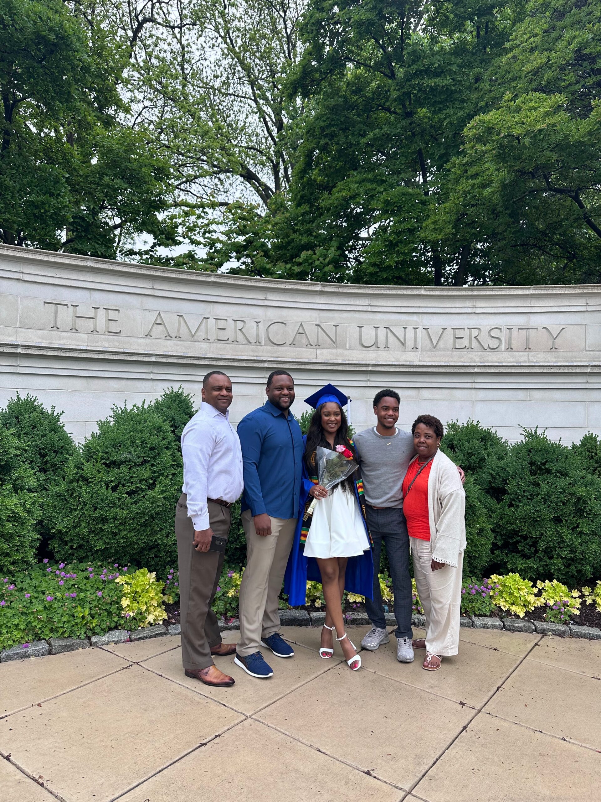Group of graduates and family posing in front of a stone wall reading 'The American University' with a graduate in a blue gown and cap holding flowers, flanked by friends and relatives outdoors with greenery.