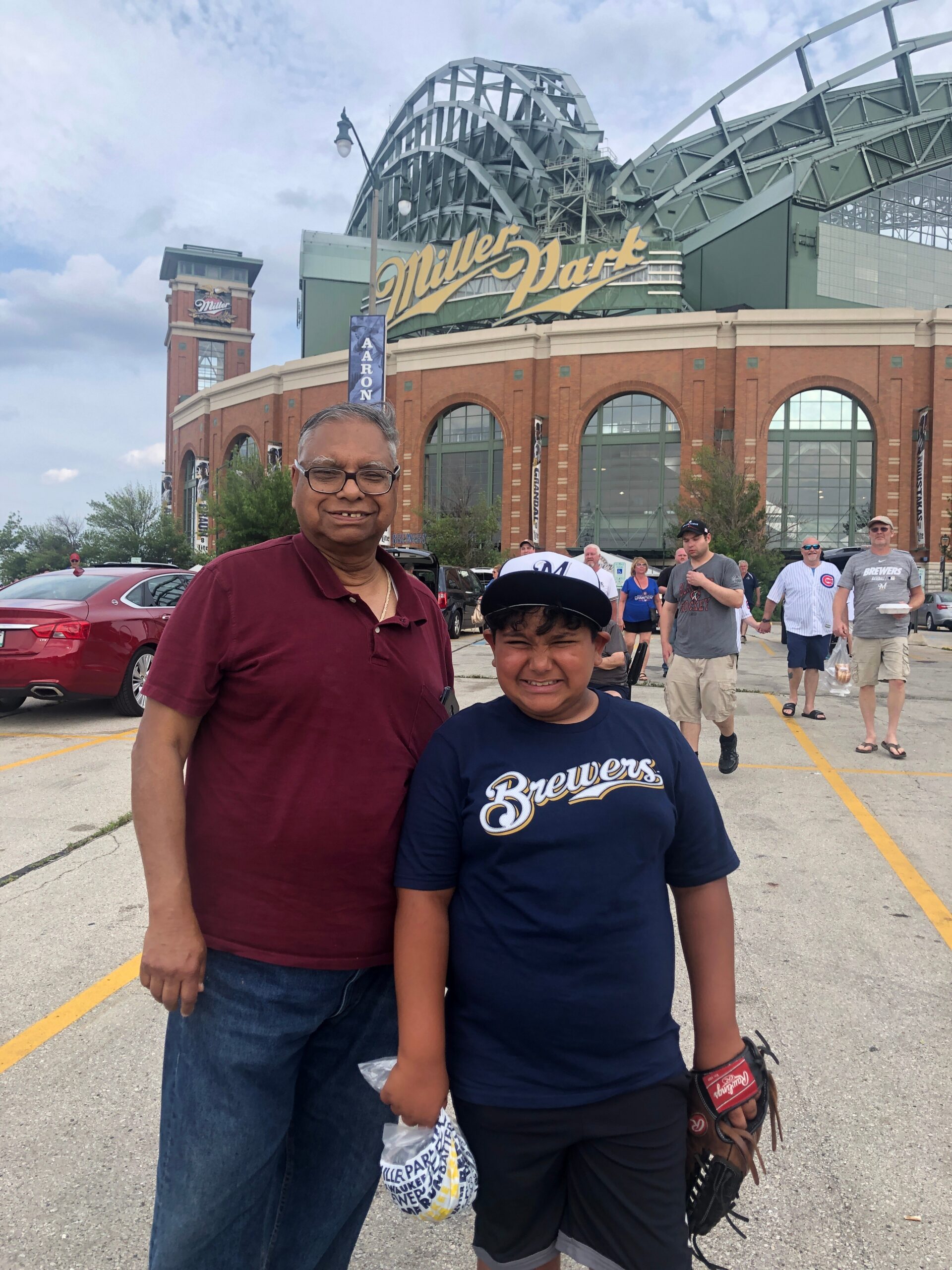 Smiling man in a maroon polo and a boy in a Brewers shirt pose in a parking lot outside Miller Park stadium, with fans in the background.