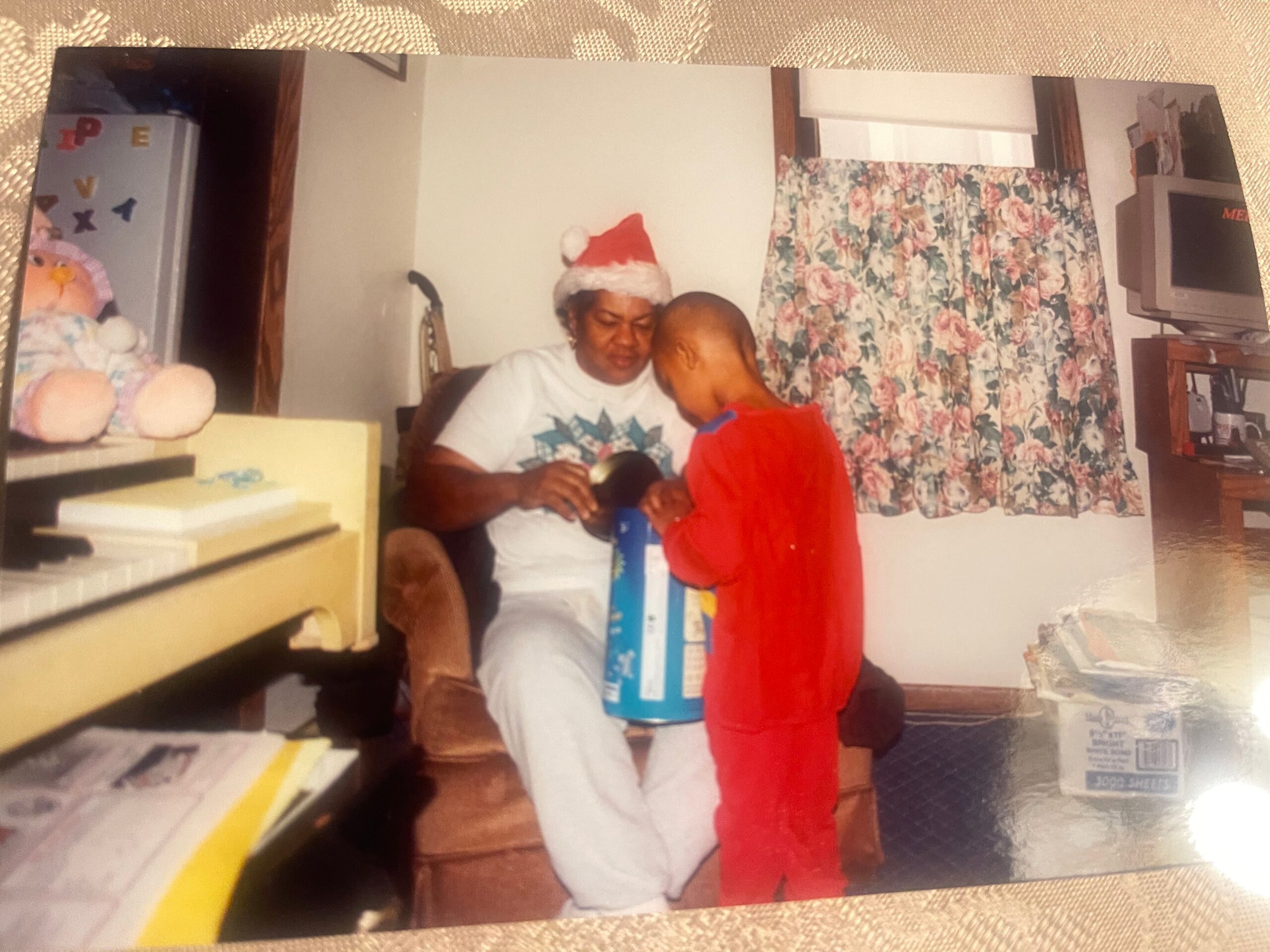 A woman wearing a Santa hat sits with a child, helping unwrap a blue gift in a cozy living room holiday scene.