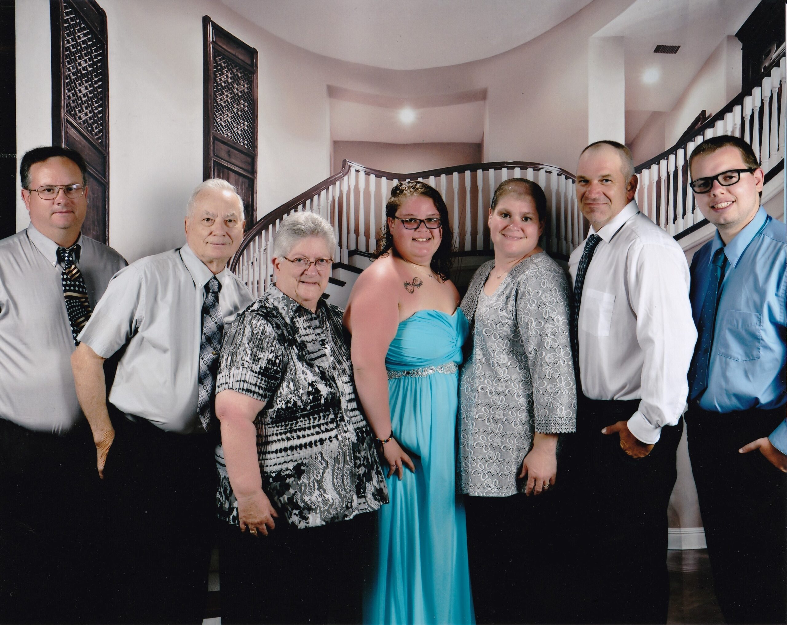Seven adults in formal attire pose together in front of a curved staircase backdrop.