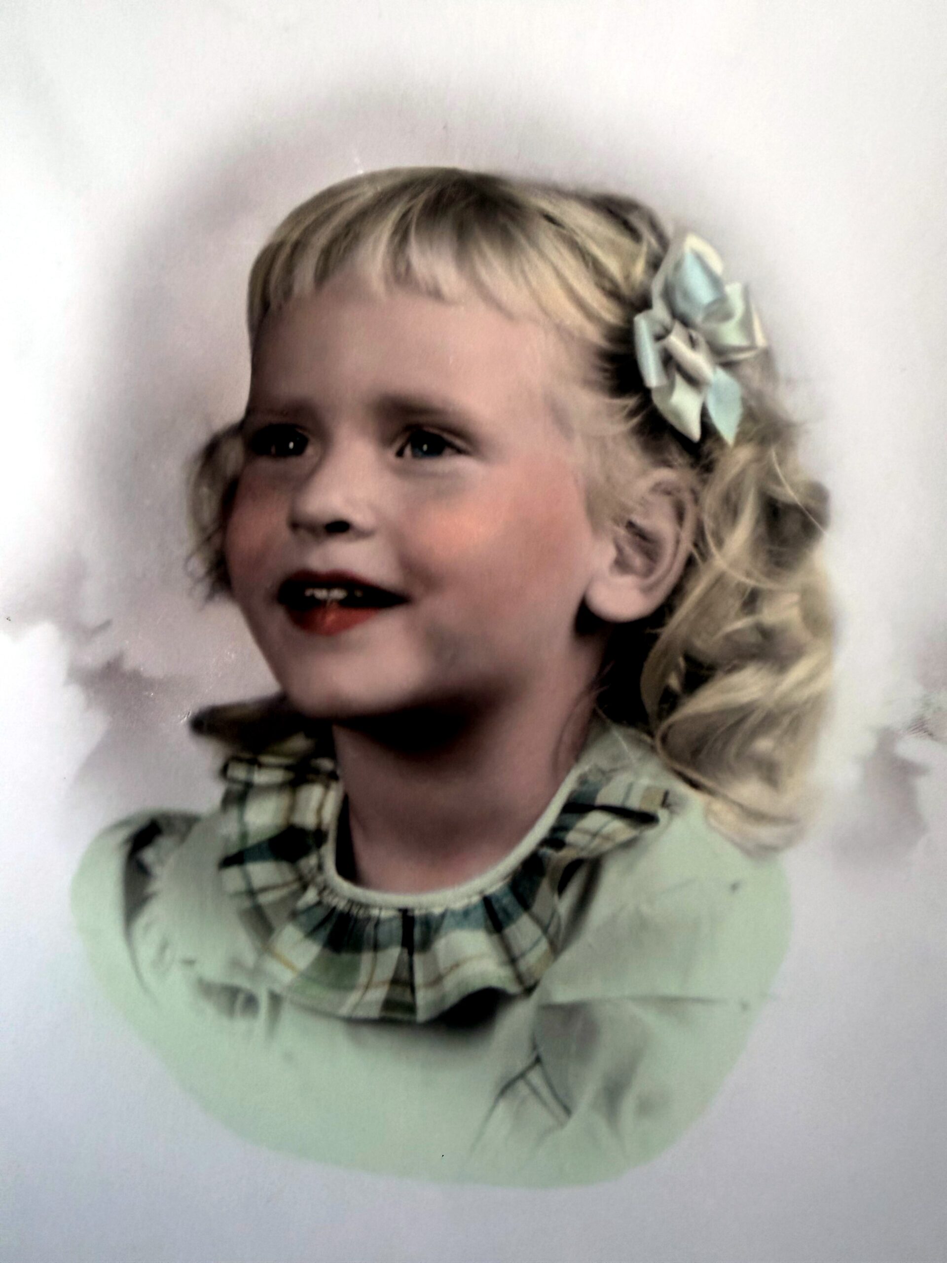 Portrait of a young girl with blonde hair in a green plaid dress, wearing a pale blue bow, smiling softly to the left.