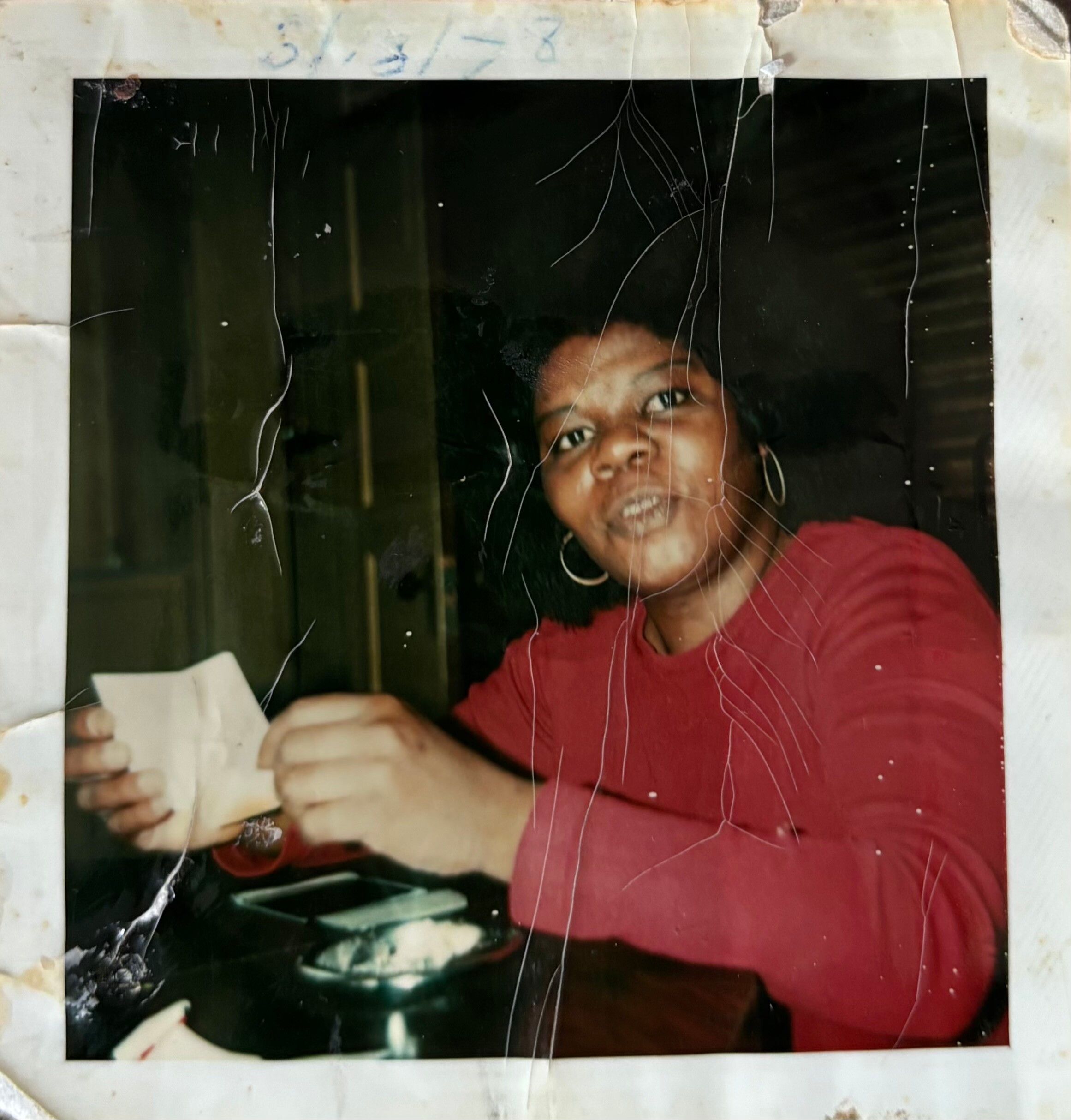 Smiling woman with hoop earrings wearing a red top, seated at a table with a plate of dessert in front.