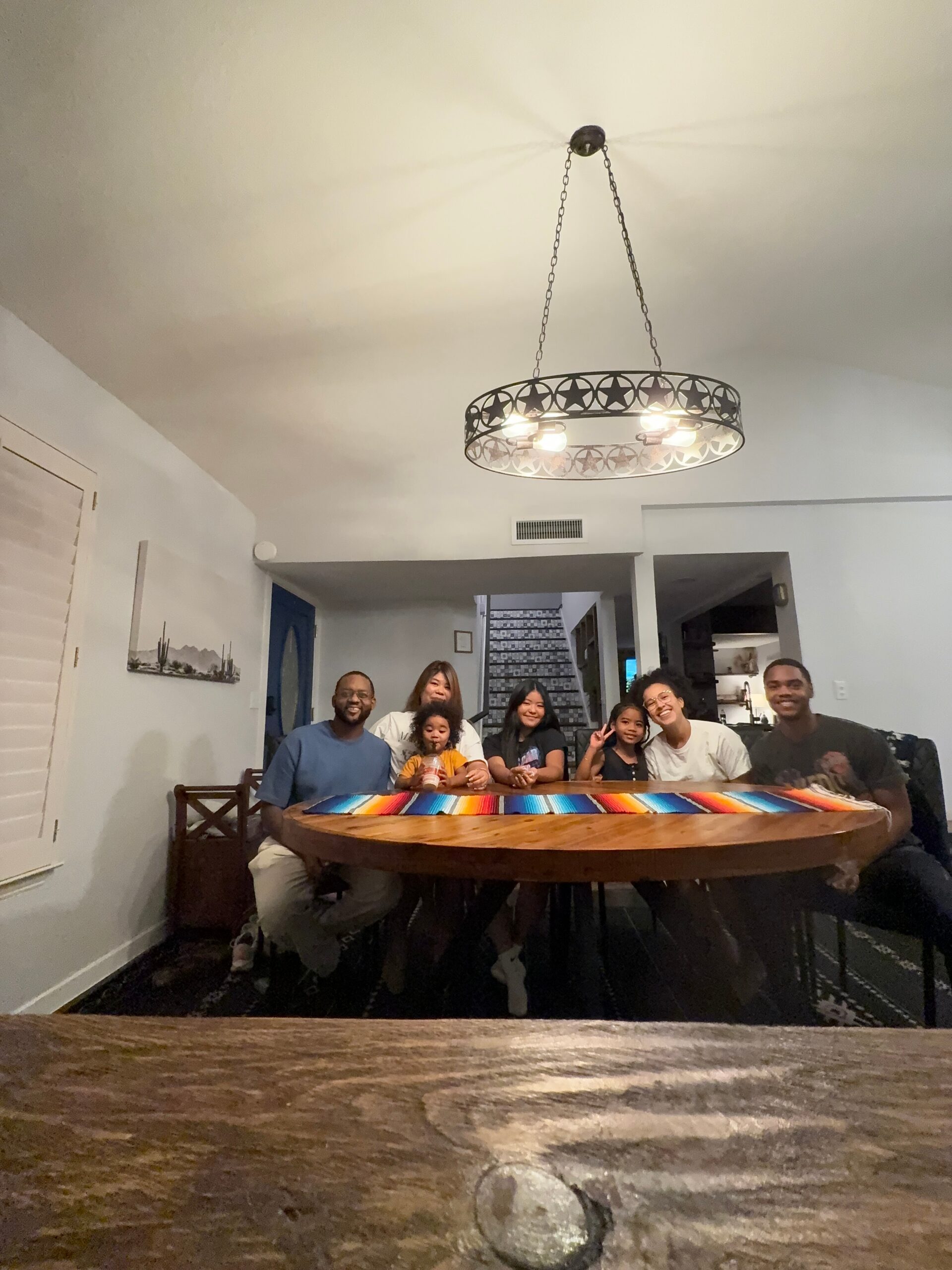 Seven people sit around a large wooden dining table with a bright striped runner, smiling at the camera in a home dining room.