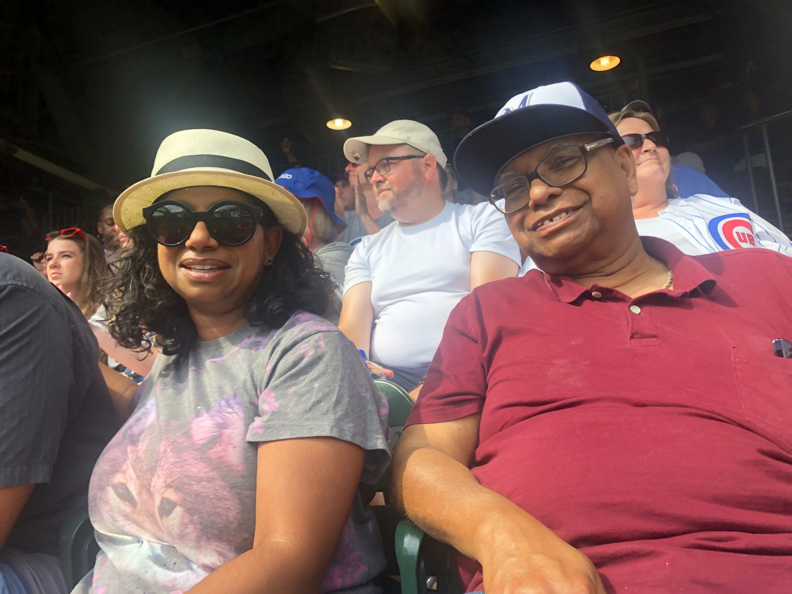 Several spectators seated in a stadium, wearing hats and sunglasses, smiling at a daytime event