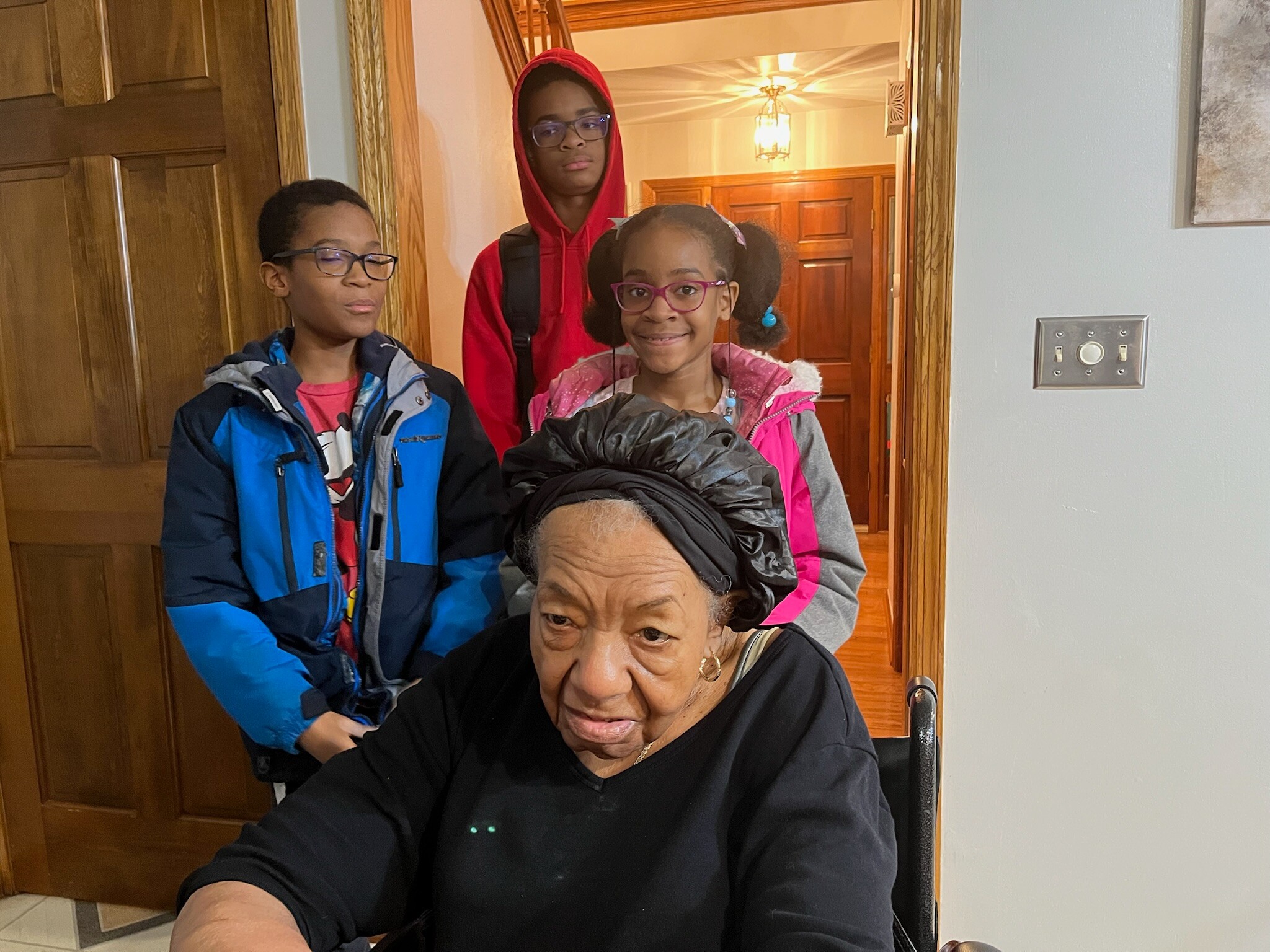Elderly woman in a wheelchair in the foreground with three children behind her in a doorway, indoors and smiling.