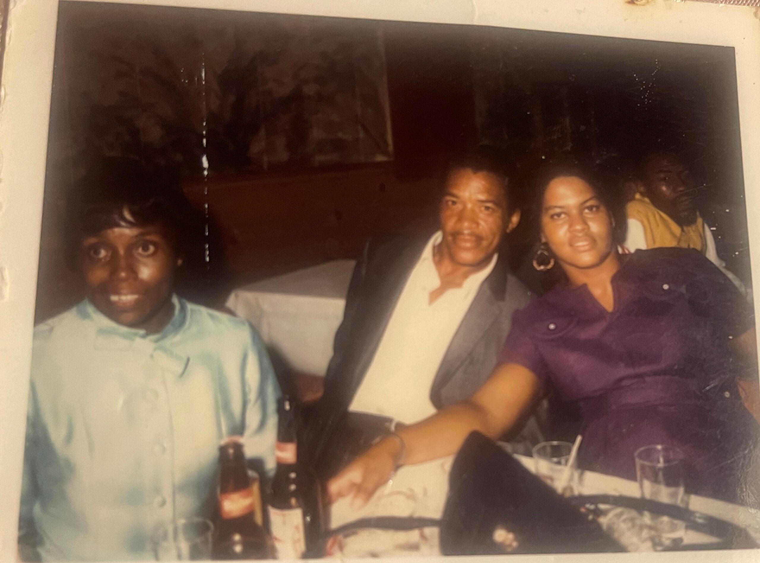 Three friends posing for a photo at a table with drinks: man in a light blue shirt on the left, man in a suit in the middle, and a woman in a purple dress on the right.