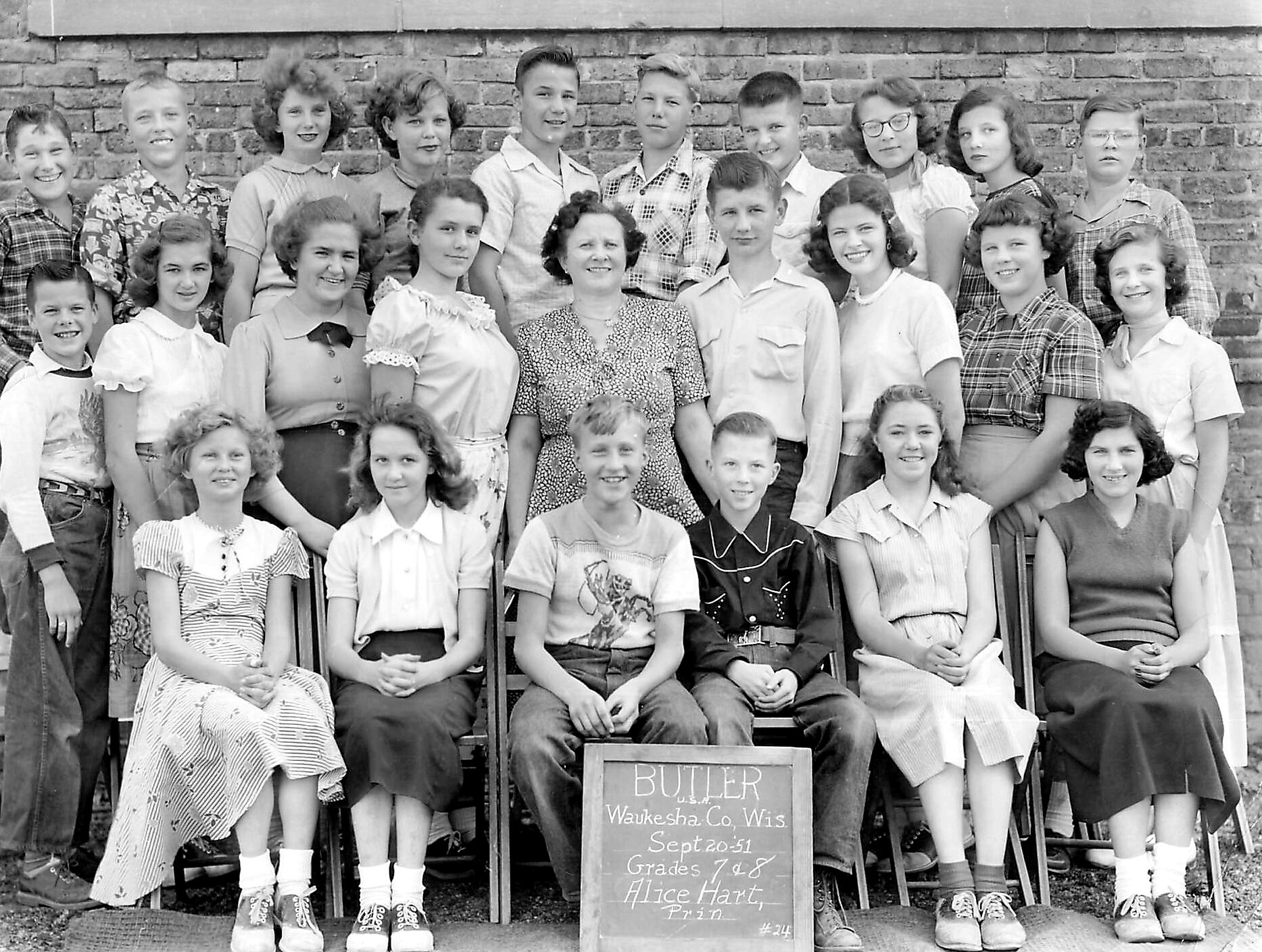 Group of schoolchildren and a female teacher posing in front of a brick wall, with a chalkboard-style sign in front listing school and class info.