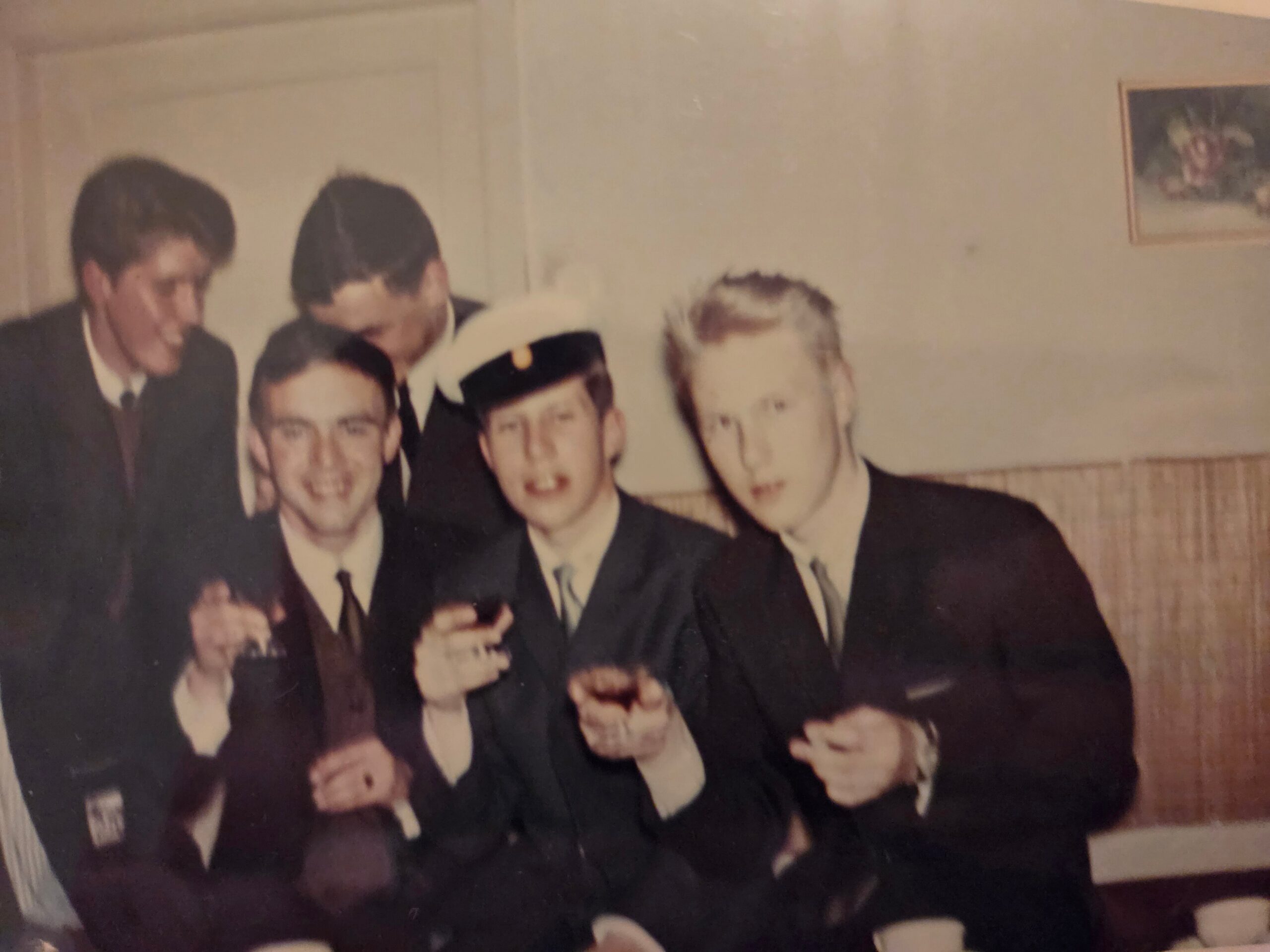 Group of people in formal suits posing together at a social gathering, some holding drinks, indoors in a room with a small framed picture on the wall.