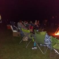 Group of people sitting in lawn chairs around a campfire at night on grass.