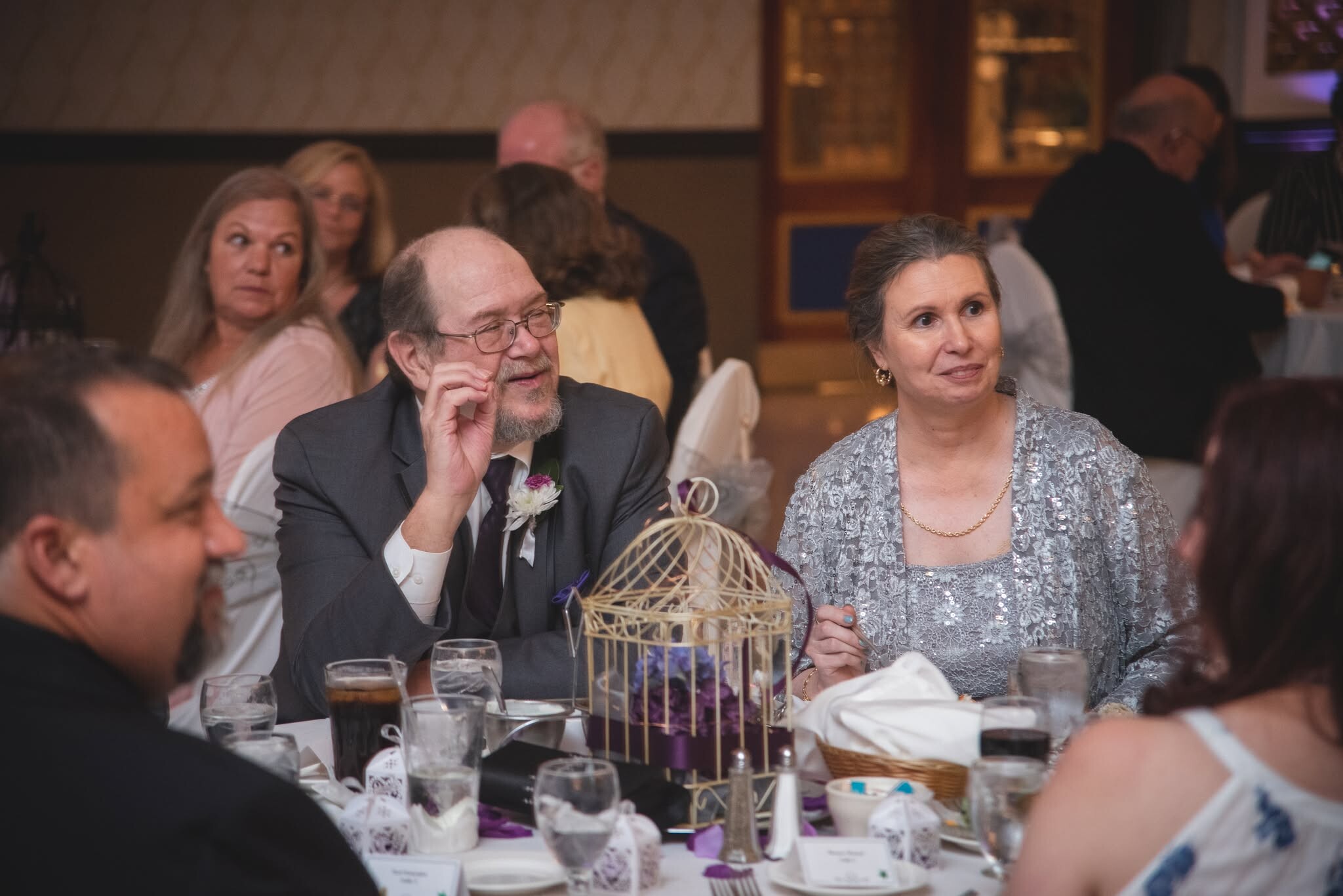 Man with glasses and a gray beard sits at a formal dinner table, talking with a woman in a silver lace dress; decorative centerpiece and place settings in foreground.