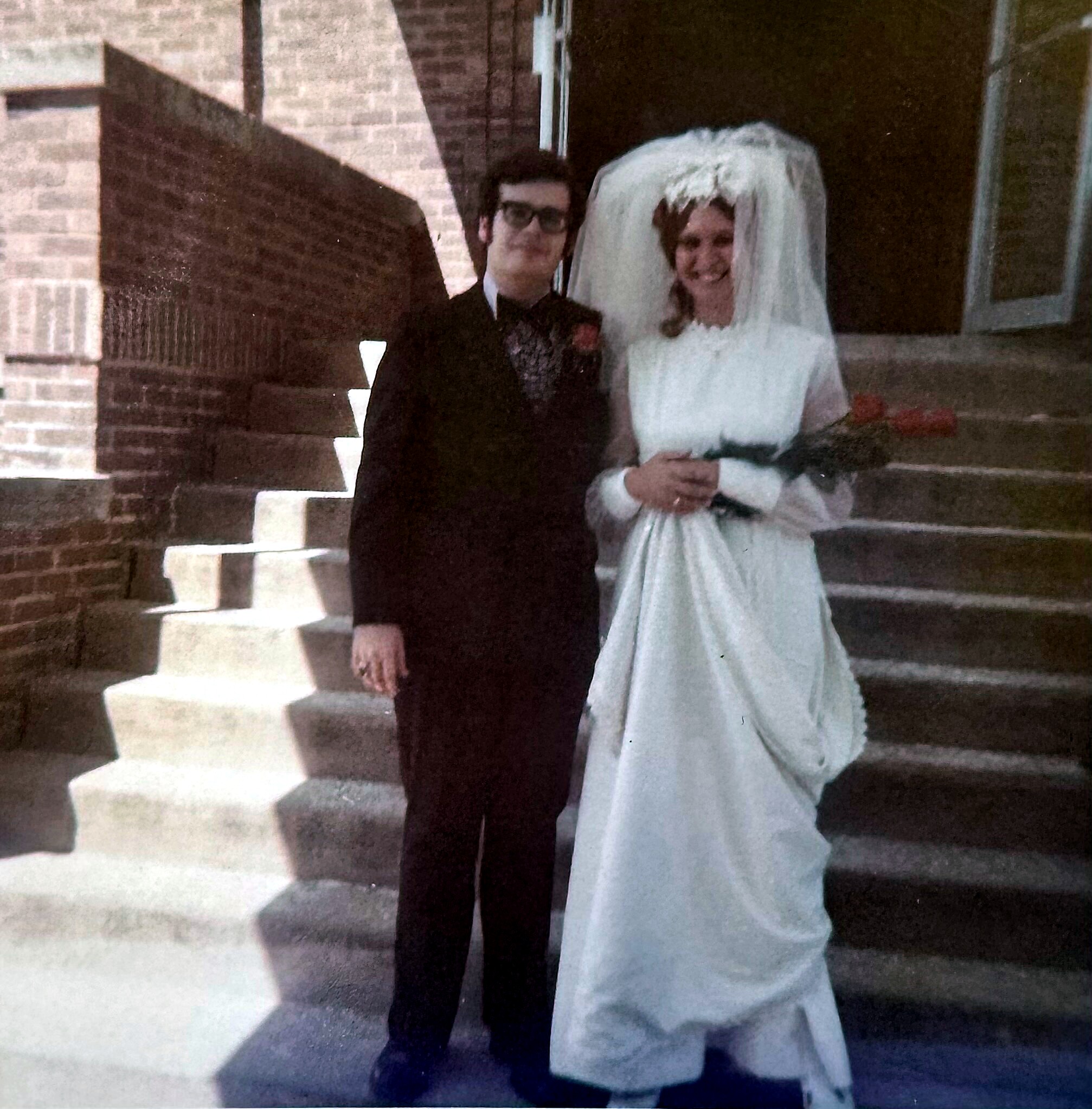 Bride in a white wedding dress and veil standing beside a man in a dark suit on stone steps outside a building.