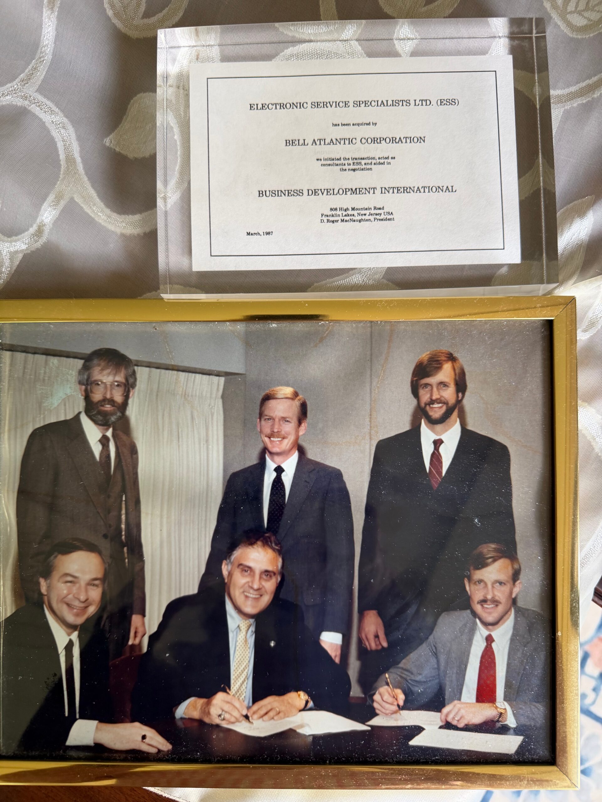 Group of men in suits signing documents at a table during a formal ceremony; framed corporate certificate above.