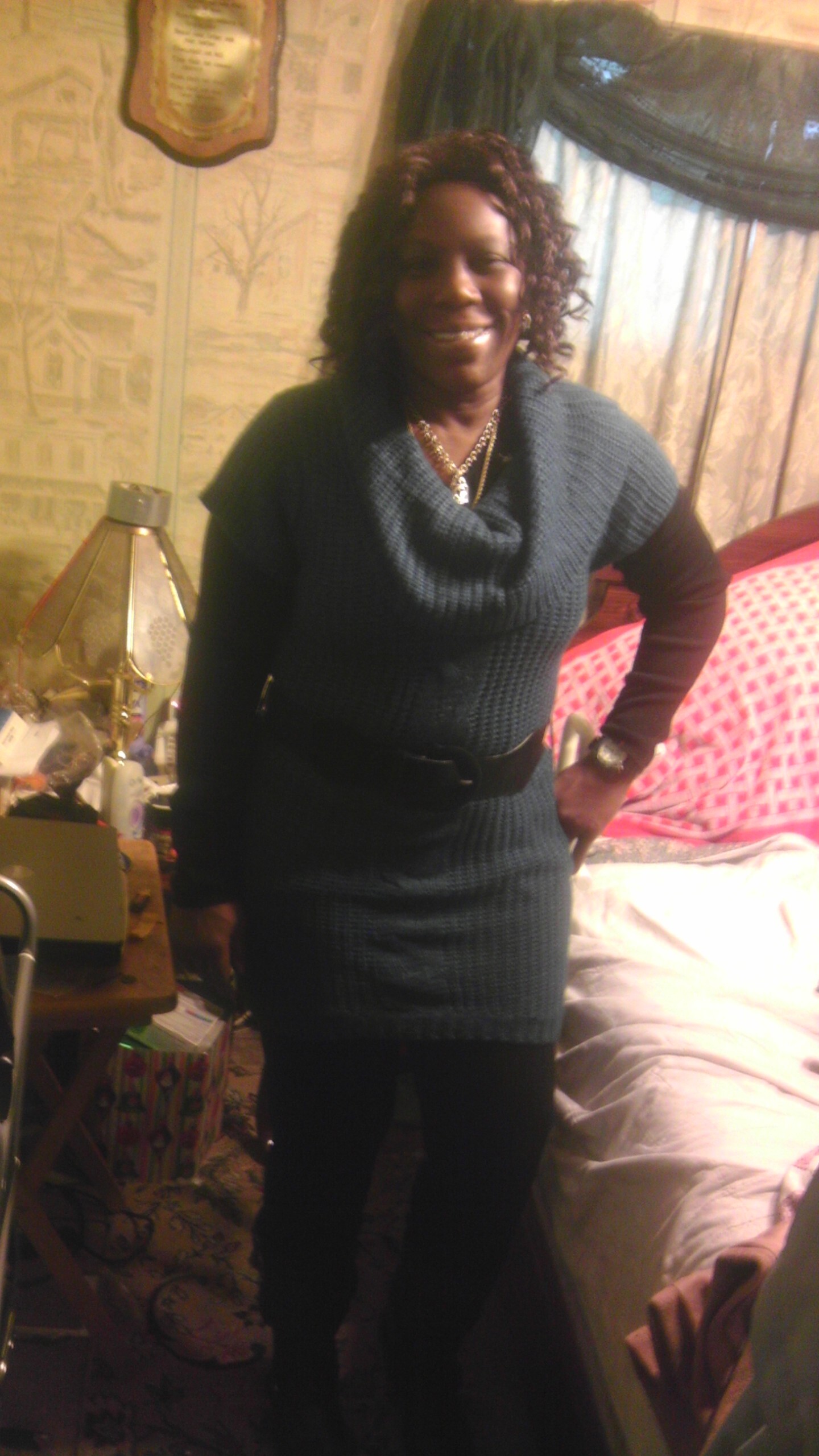 Smiling woman standing in a cluttered bedroom wearing a teal knit dress with a belt.