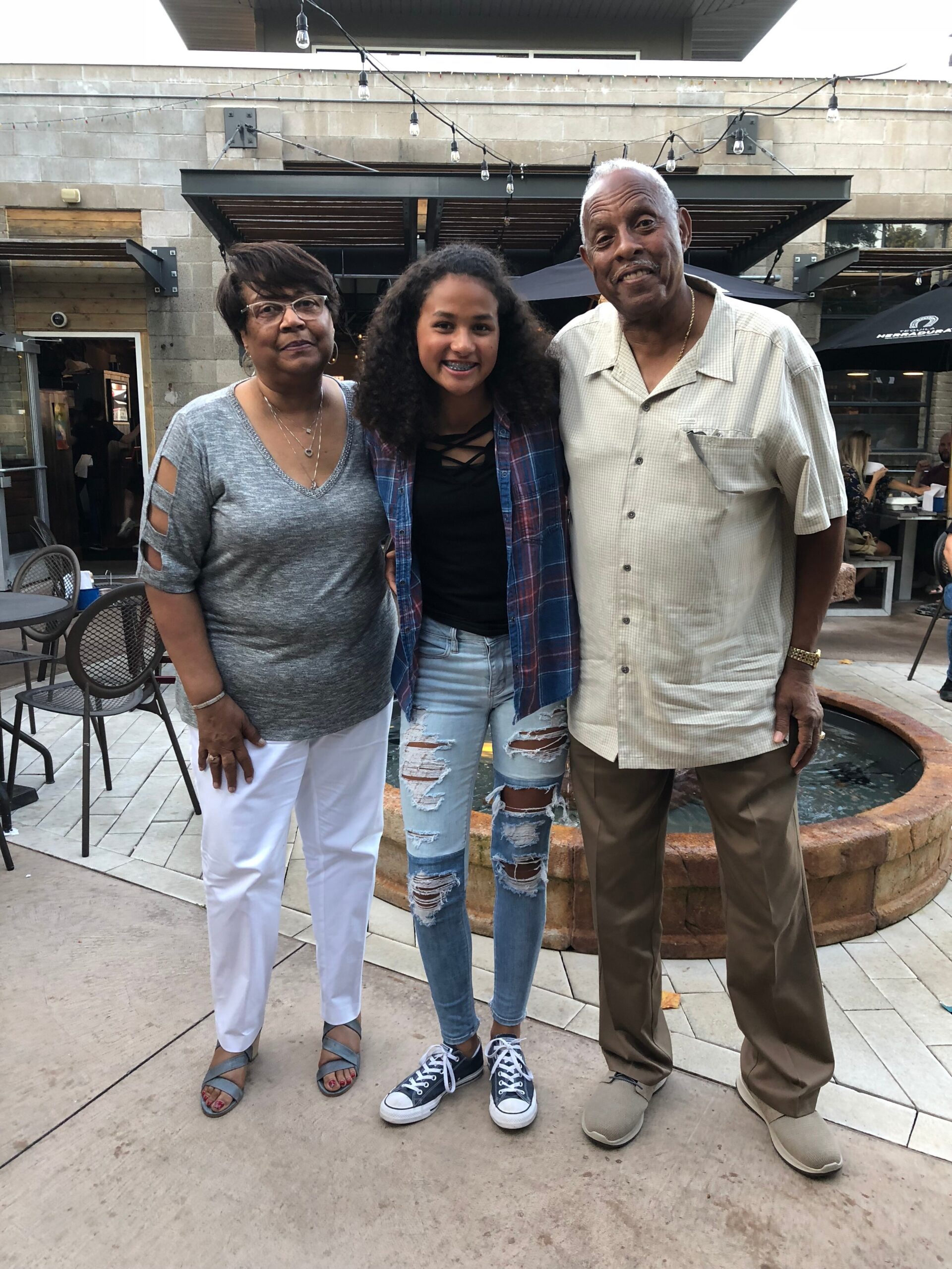 Three generations posing together outdoors near a fountain: an older woman in gray top and white pants, a teenage girl in a plaid shirt and ripped jeans, and an older man in a beige short-sleeve shirt.