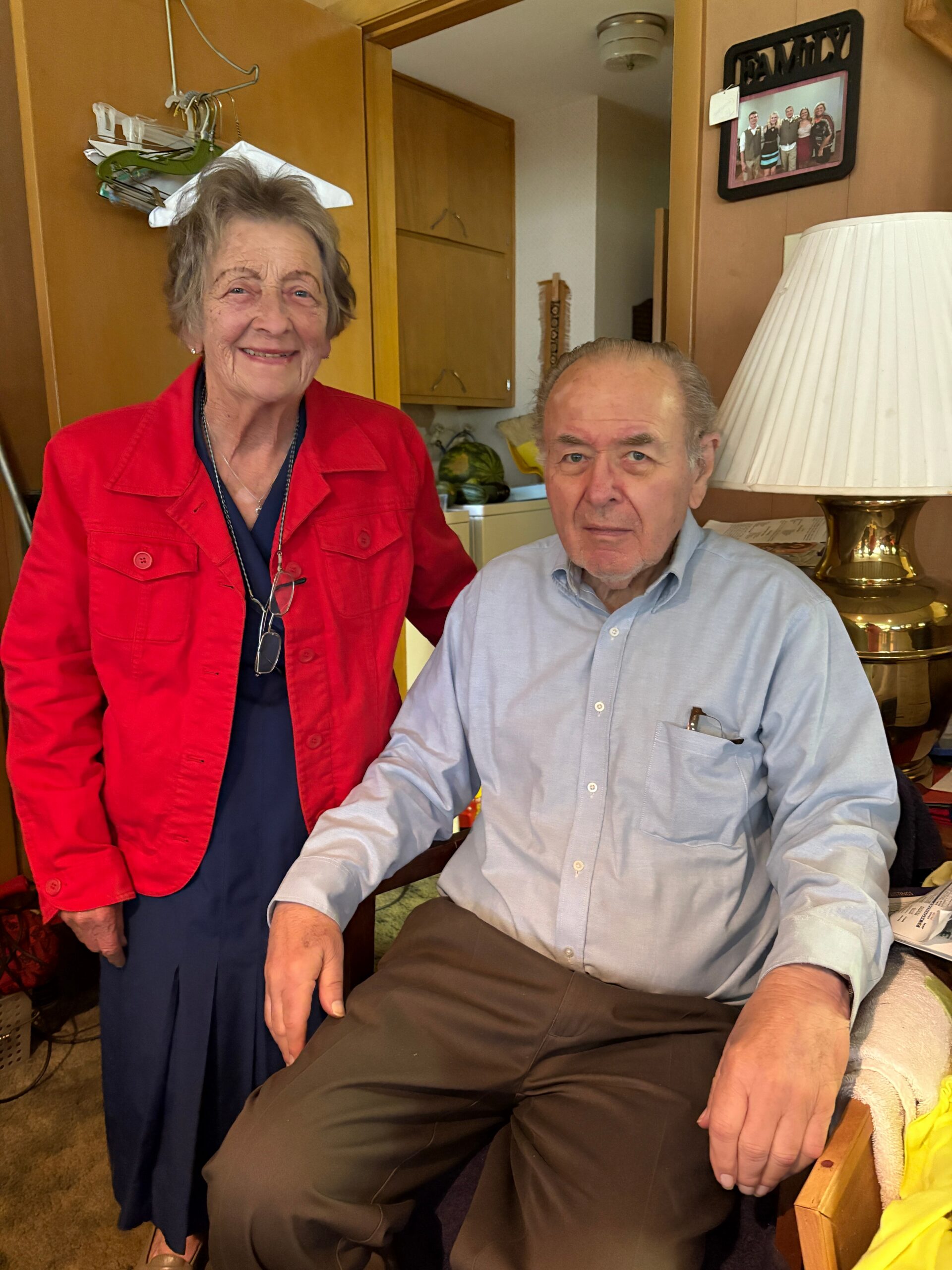 Smiling elderly woman in a red jacket stands beside a seated older man in a light blue shirt in a cozy living room with wooden cabinets and a brass lamp behind them.