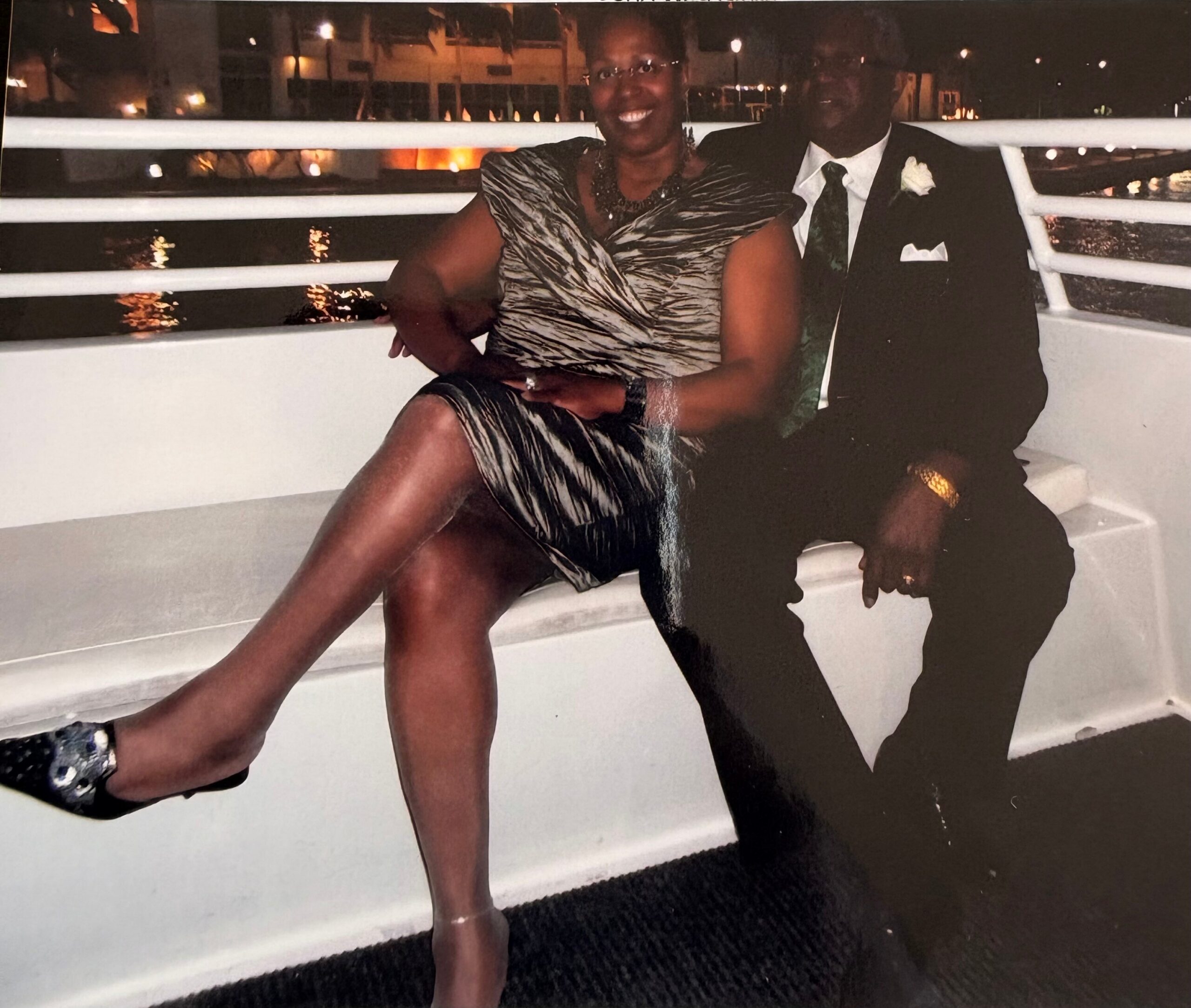 Couple sitting on a boat railing at night, smiling, dressed formally in a patterned dress and suit.