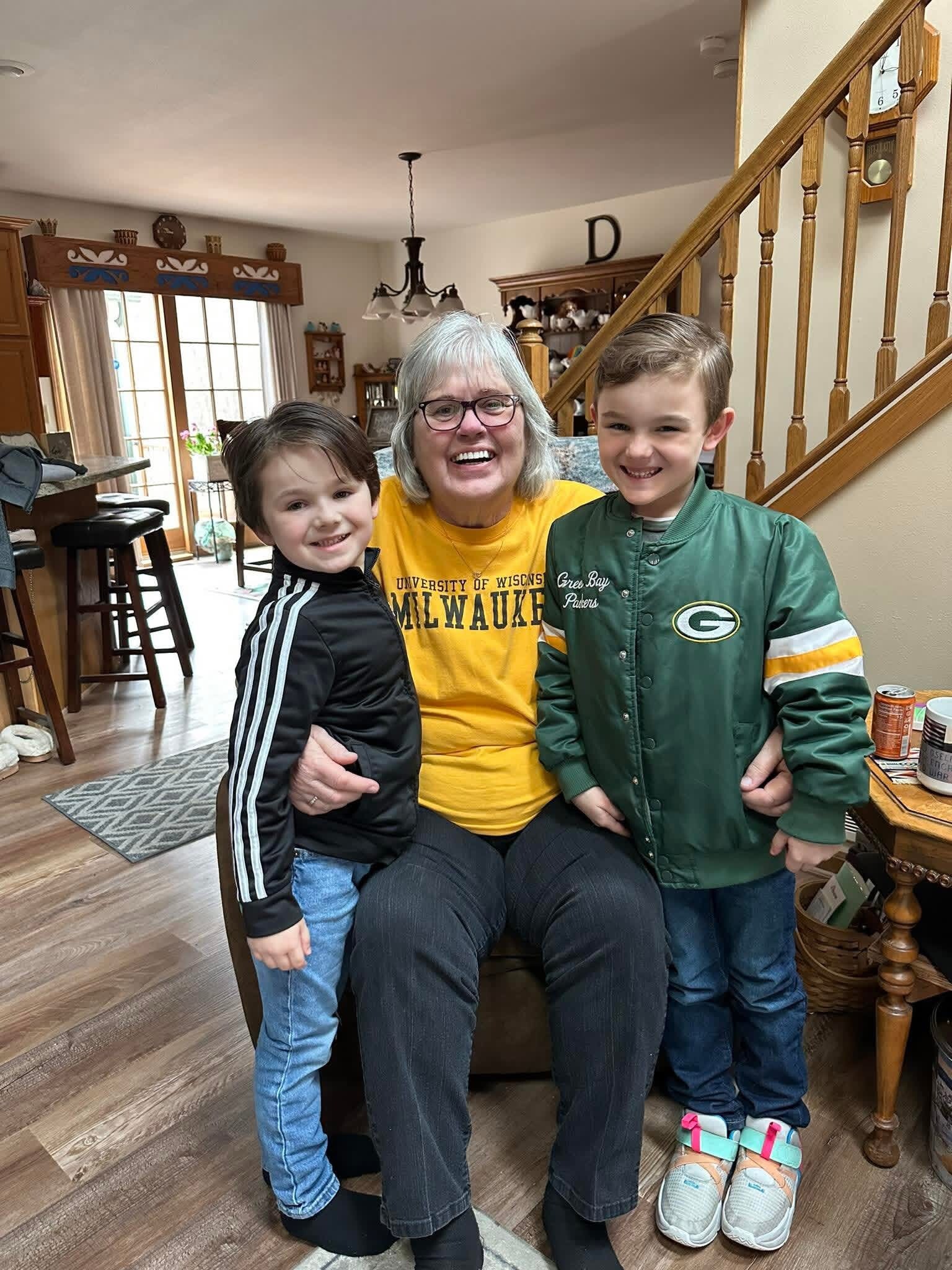 Grandmother with two young boys posing and smiling in a cozy living room with a wooden staircase behind them.