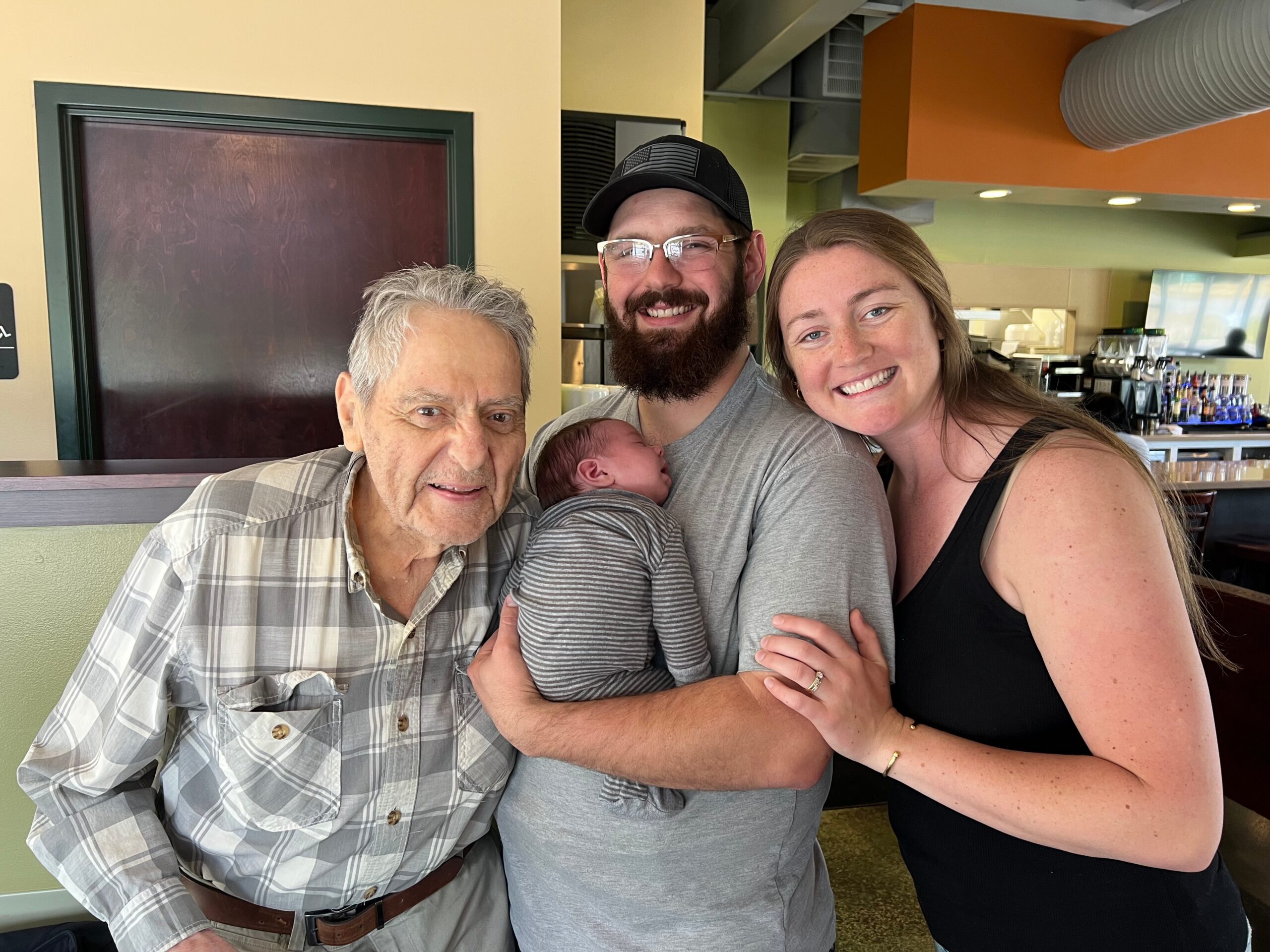 Smiling three‑generation family posing in a cafe: grandfather on the left, a bearded man holding a newborn baby, and a woman on the right.