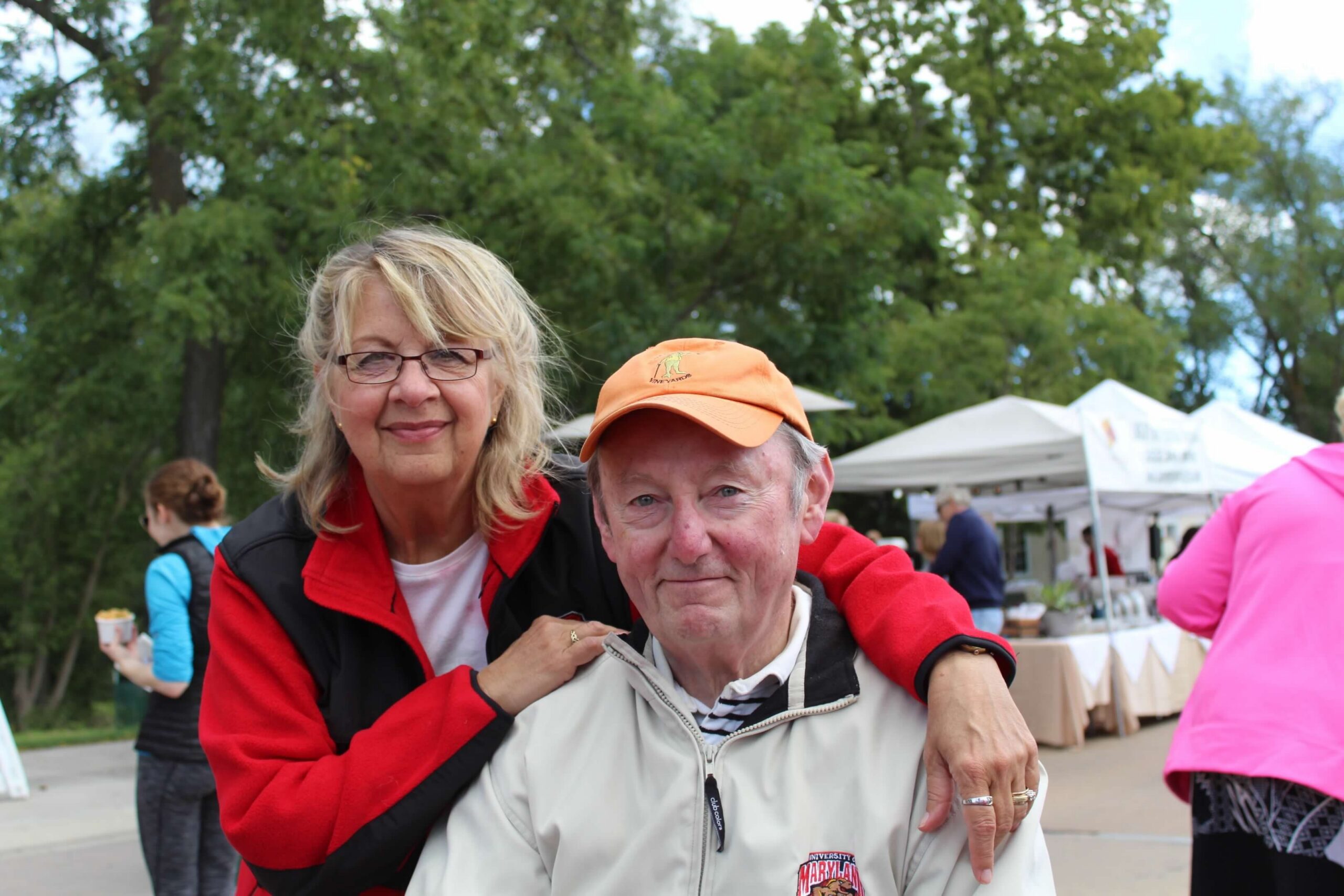 Two smiling adults pose together at an outdoor market or festival, with trees and white vendor tents in the background.