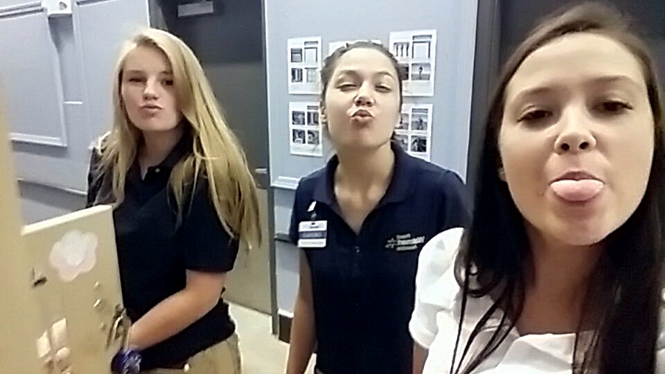 Three women stand in a hallway, posing and making funny faces at the camera. Two wear dark polo shirts; one wears a white blouse with long hair showing on the left side.