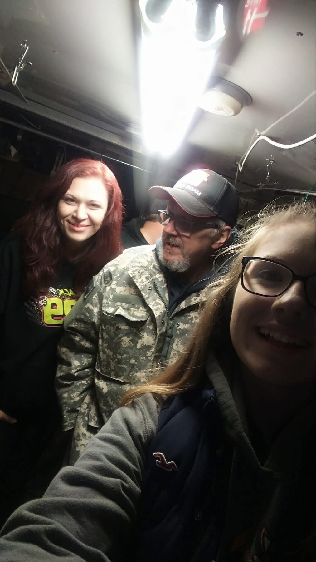 Three friends pose for a selfie in a dim, metal interior: red-haired woman on the left, a man in camouflage jacket and cap center, and a girl with glasses on the right.