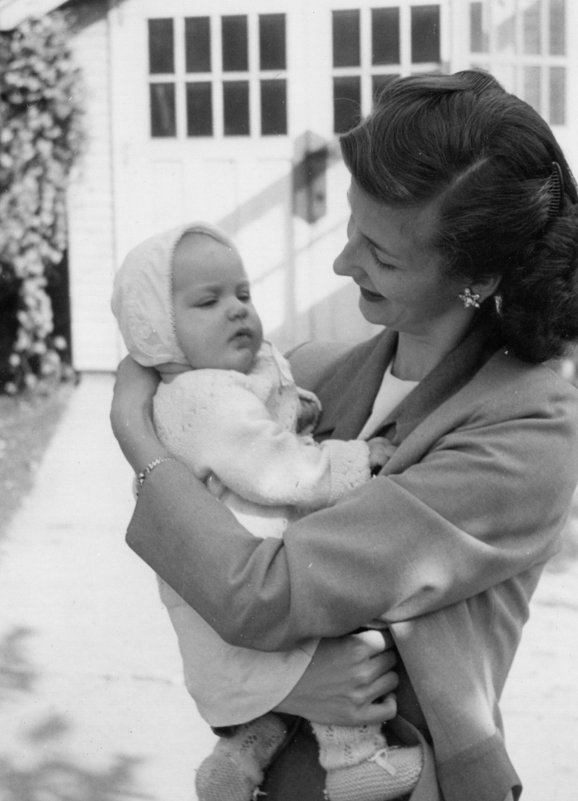 Woman in a blazer smiling while holding a baby in a knit bonnet outside a house with windows in the background