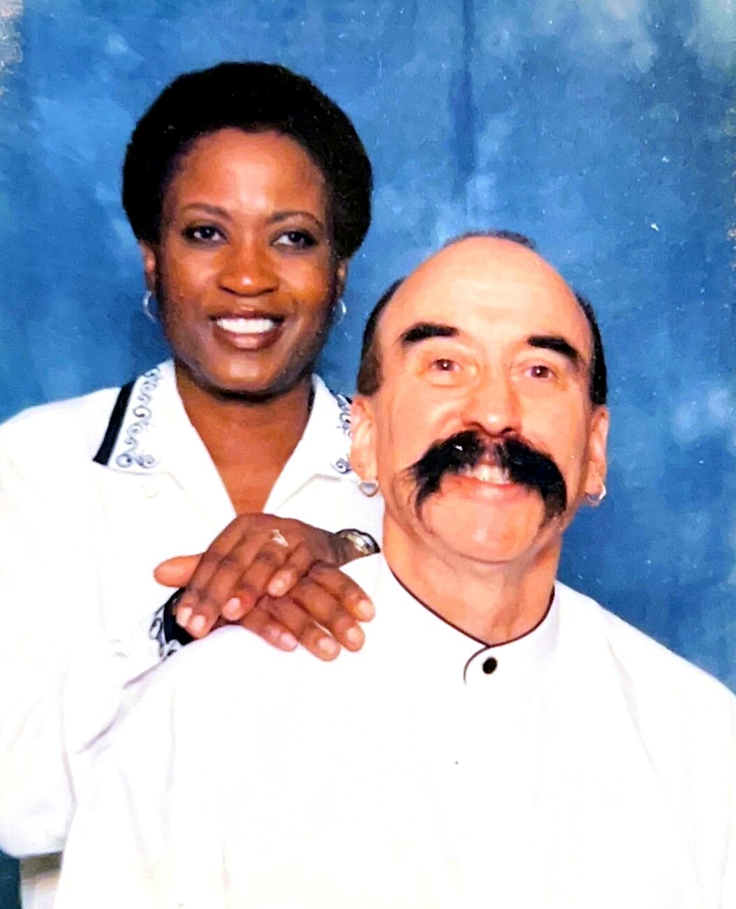 Smiling Black woman stands behind a man with a thick mustache, both in white, against a blue backdrop.