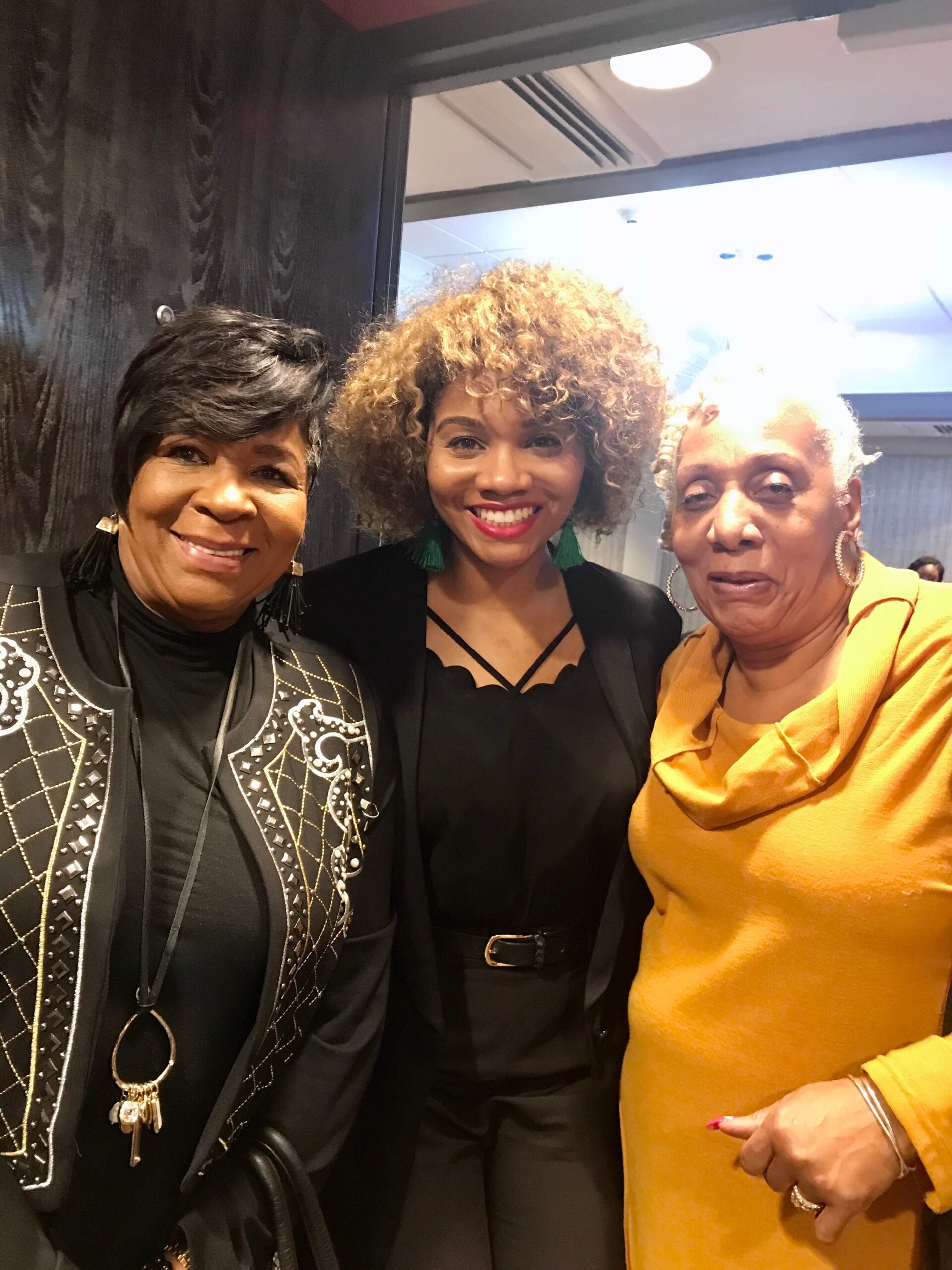 Three women posing for a close-up selfie indoors, all smiling; left and center in black outfits, right in a mustard-yellow top.
