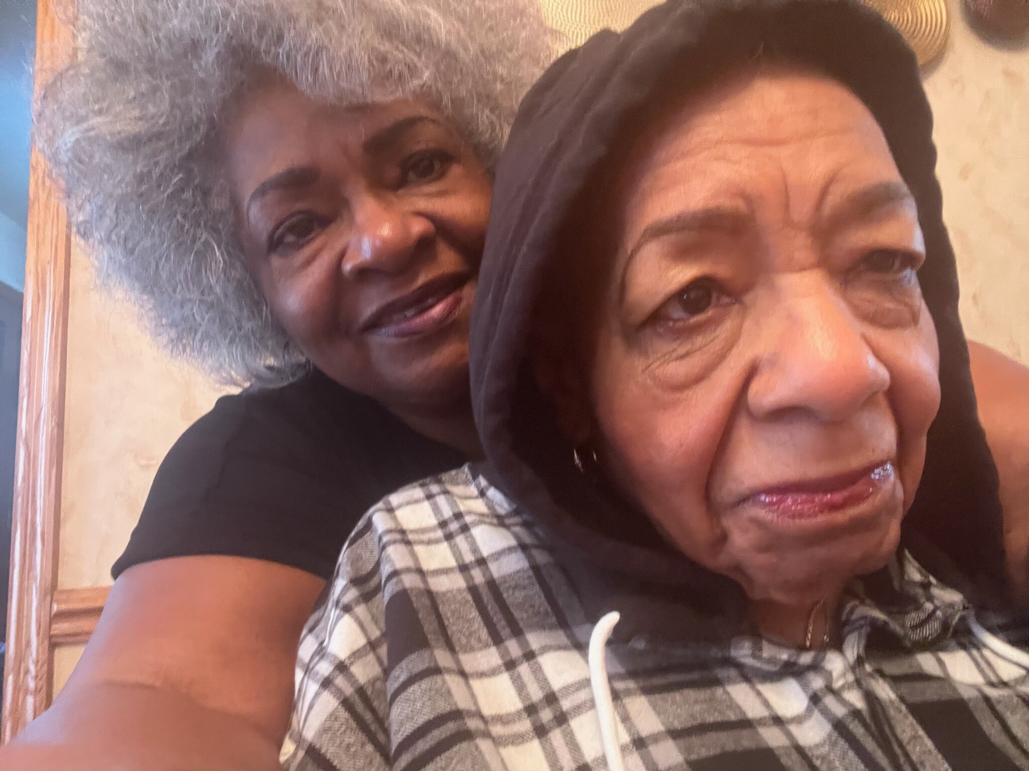 Two women posing closely for a selfie indoors, smiling at the camera; one with gray curly hair on the left and a younger woman wearing a dark scarf on the right.