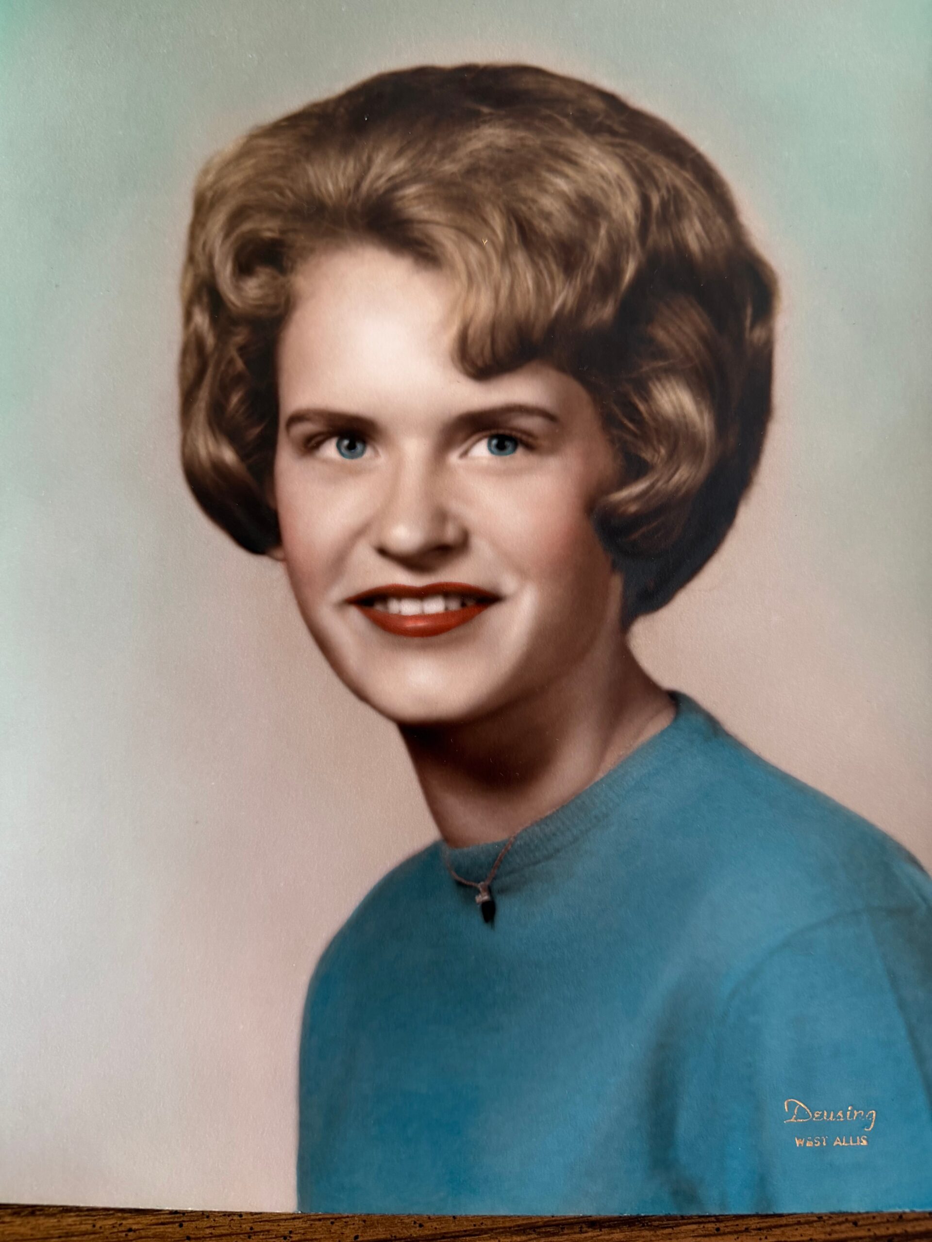 Portrait of a young woman with short blonde hair, blue eyes, wearing a teal sweater and a delicate necklace against a soft backdrop.