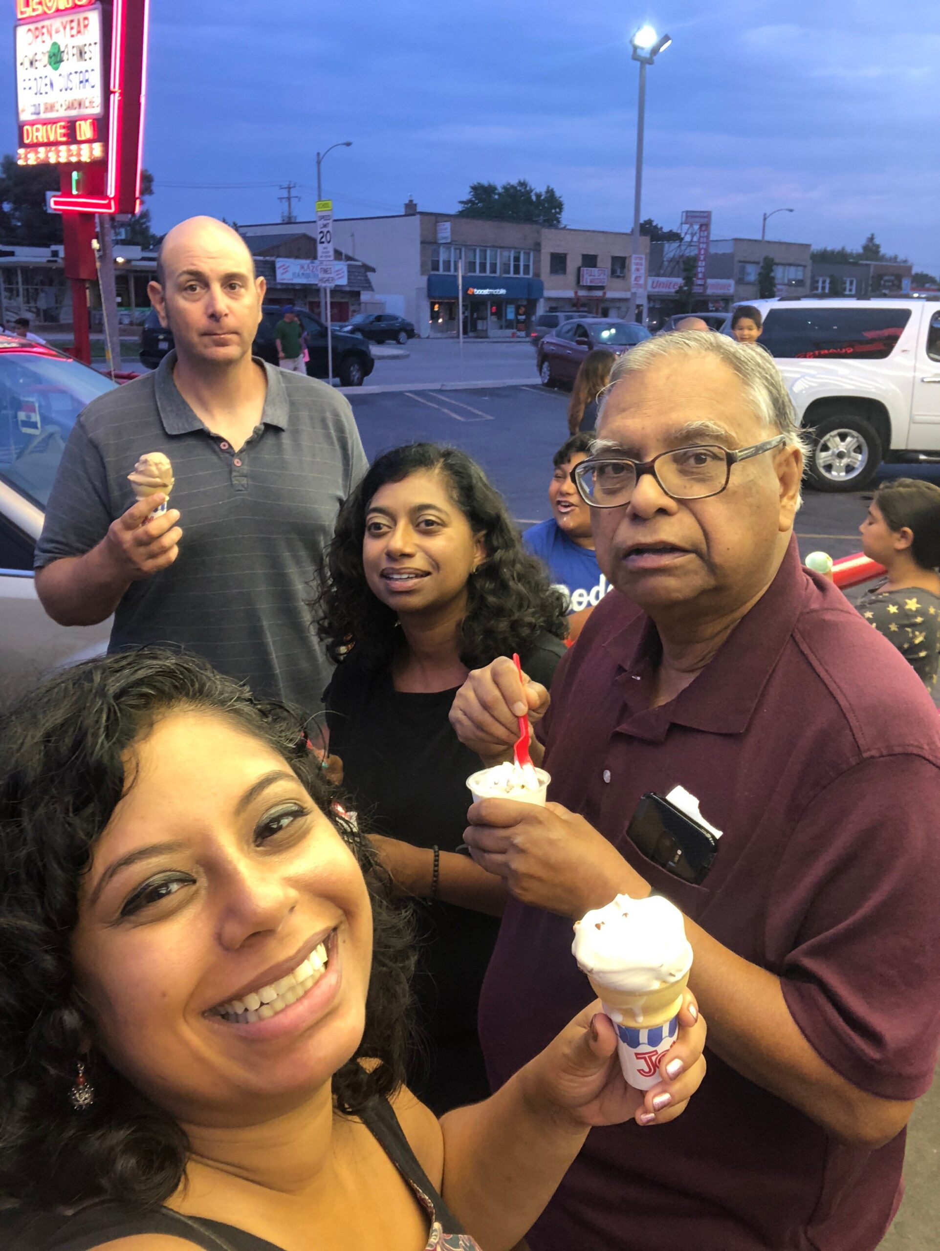 Smiling group of people posing for a selfie with ice cream cones in a parking lot, storefronts in the evening background.
