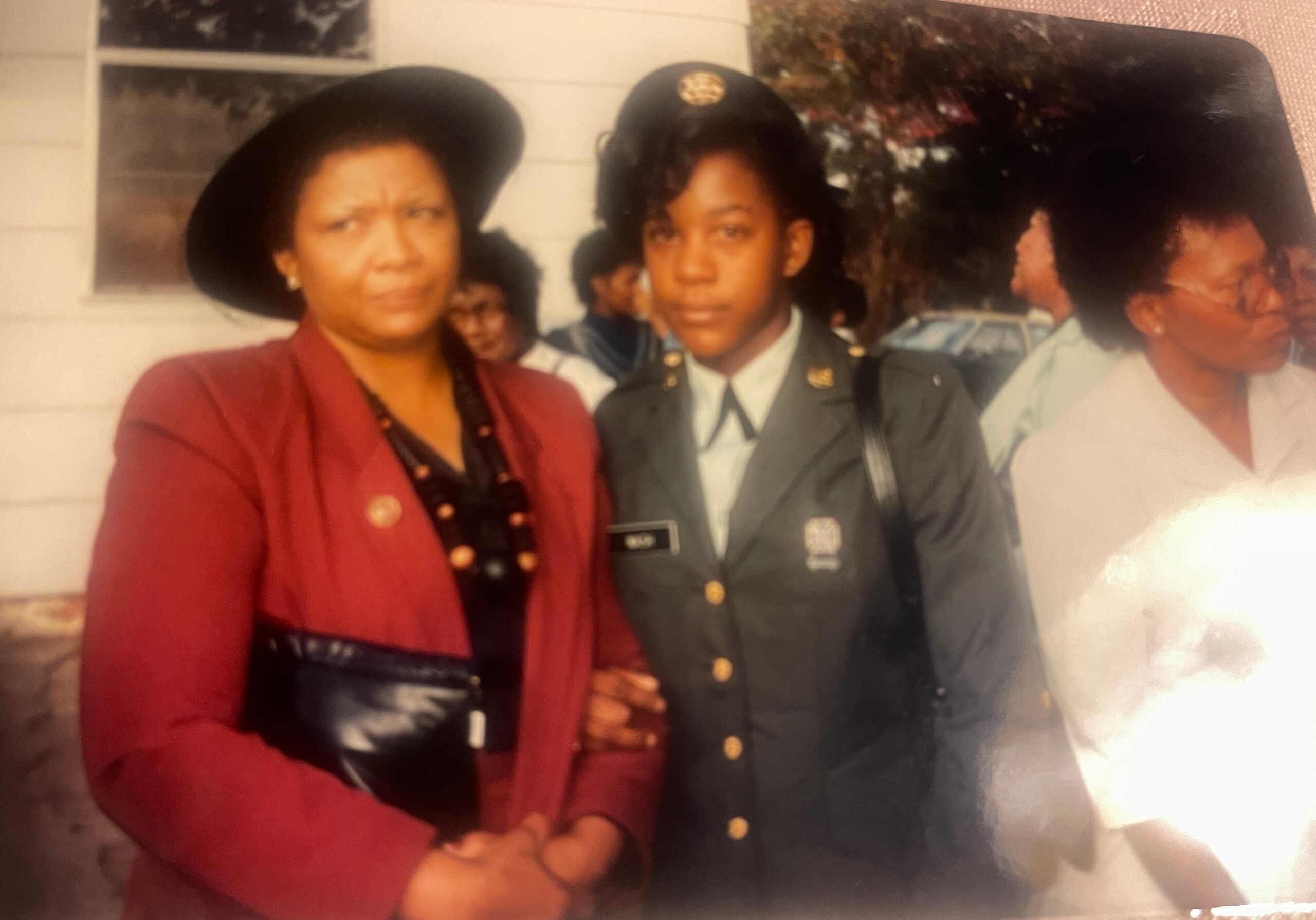 Woman in a red blazer posing with a young man in a military dress uniform, with others partially visible in the background.