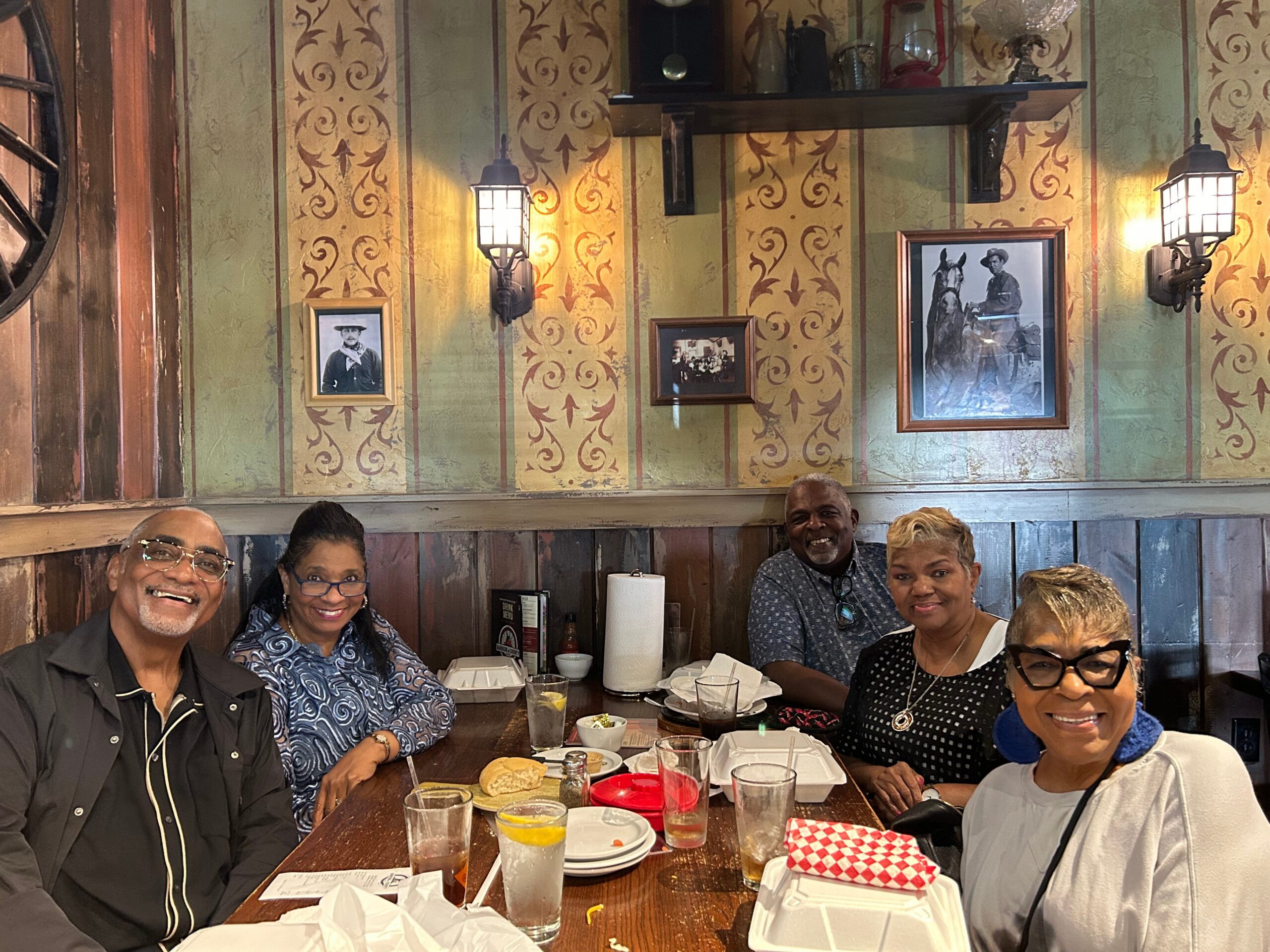 A group of six people seated around a dining table in a rustic restaurant, smiling at the camera with drinks, bread, and plates in front of them against a decorative wallpaper wall.