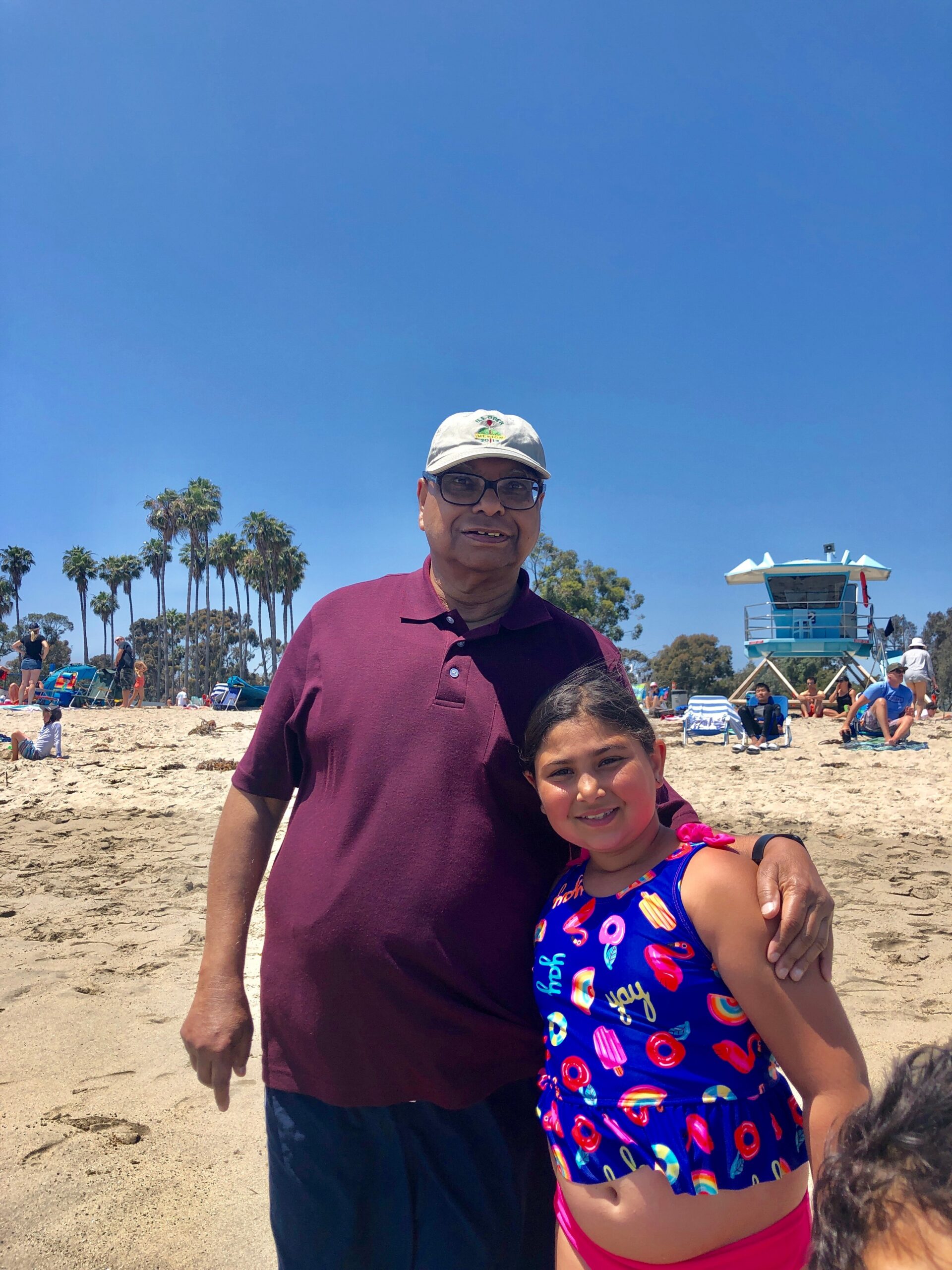 Man and young girl posing together on a sunny beach, with palm trees and a lifeguard tower in the background.