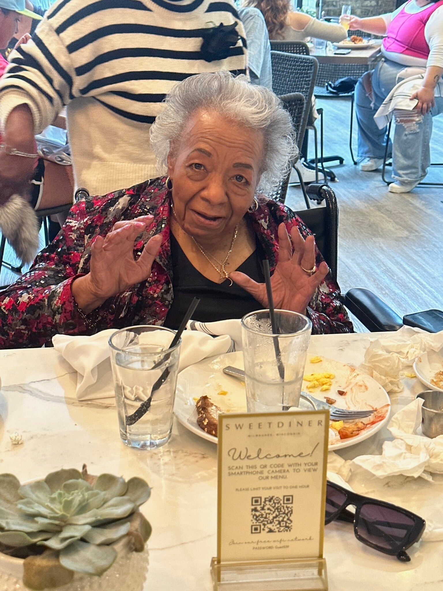 Older woman in a wheelchair at a restaurant table, raising hands in a friendly gesture with plates and drinks in front of her.