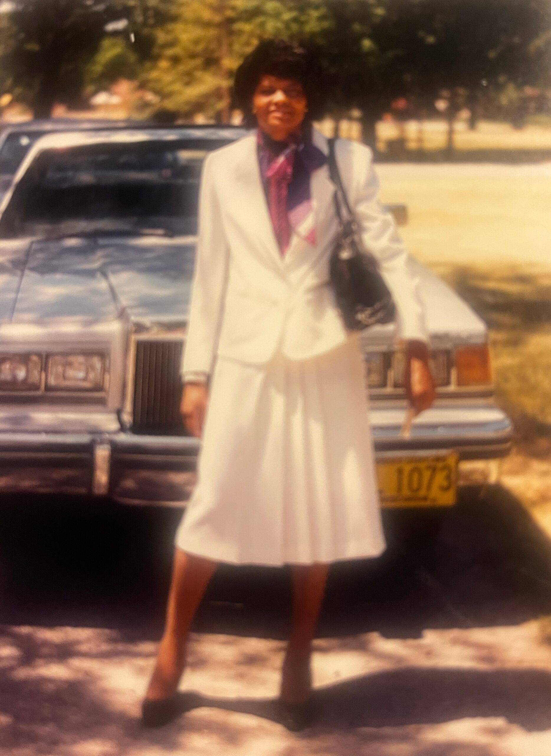 Person wearing a light-colored suit and scarf stands in front of a car on a sunny day, smiling toward the camera.
